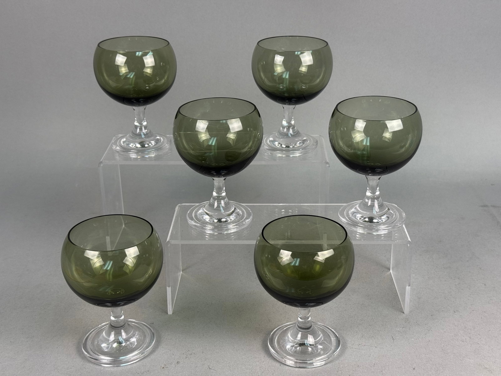 Fostoria Smoke Green Glass Goblets: A set of six mid-century modern glass goblets featuring rounded balloon-shaped bowls in a translucent smoke green hue. The colored bowls are set on short, tapered clear glass stems and circular bases.