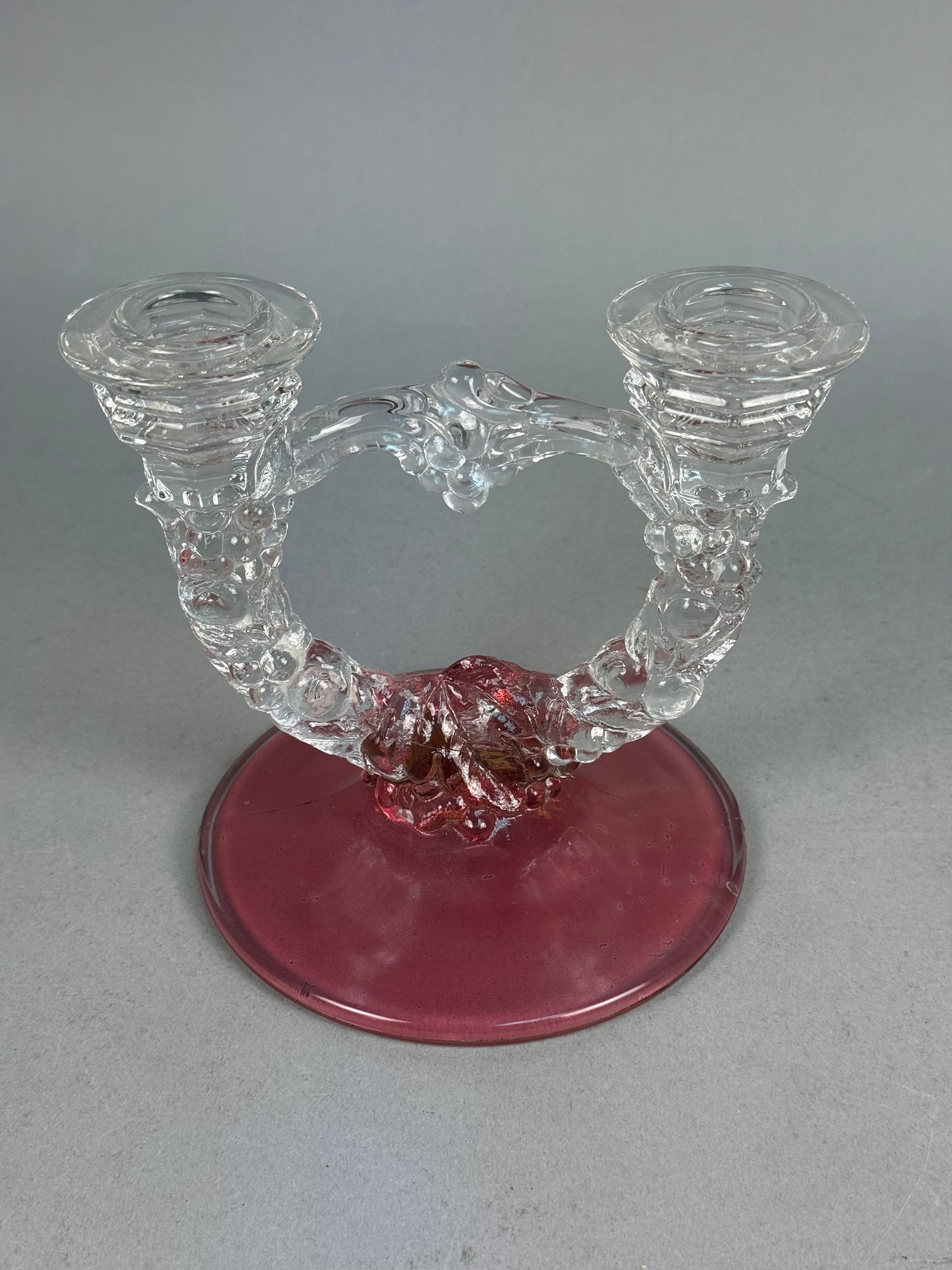 Ruby Red & Clear Glass Double-Arm Candelabrum: A vintage double-arm glass candelabrum featuring a molded grape garland motif. The piece is constructed with a circular, domed base and a central stem in a deep ruby red tone, while the arched arms an