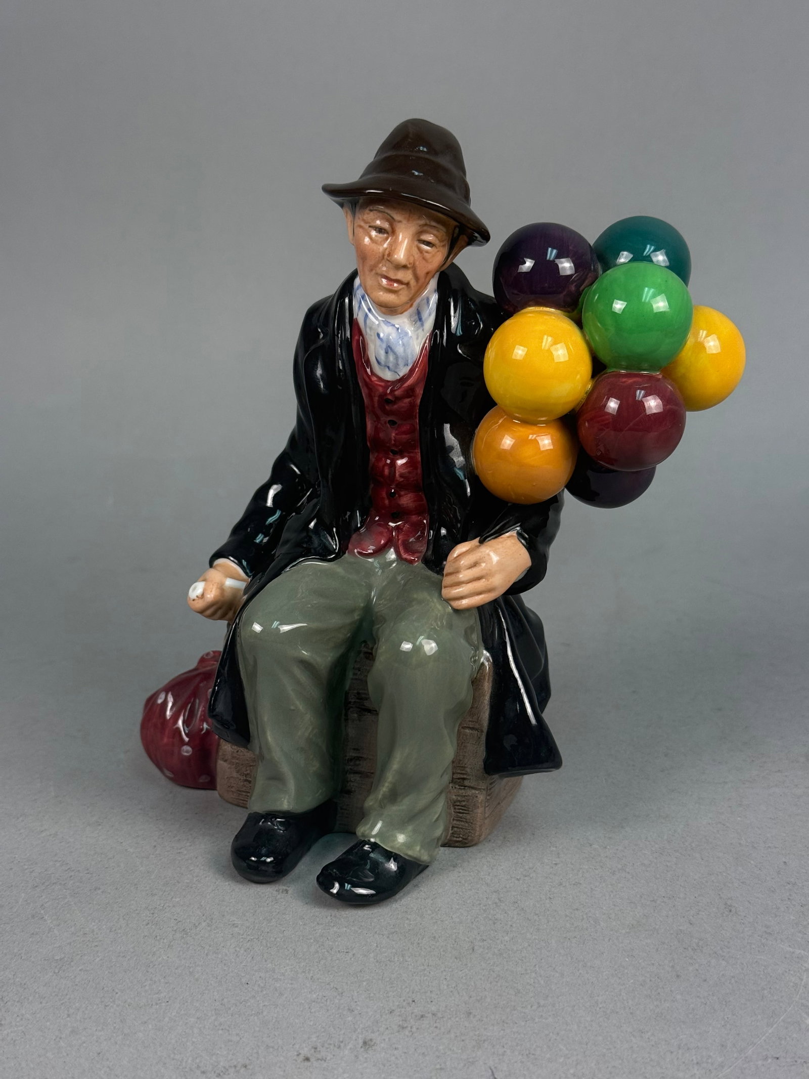 Royal Doulton Porcelain Balloon Man Figurine: This polychrome glazed porcelain character figurine by Royal Doulton is titled "The Balloon Man." The piece depicts an elderly street vendor seated upon a wooden crate, clutching a vibrant cluster of