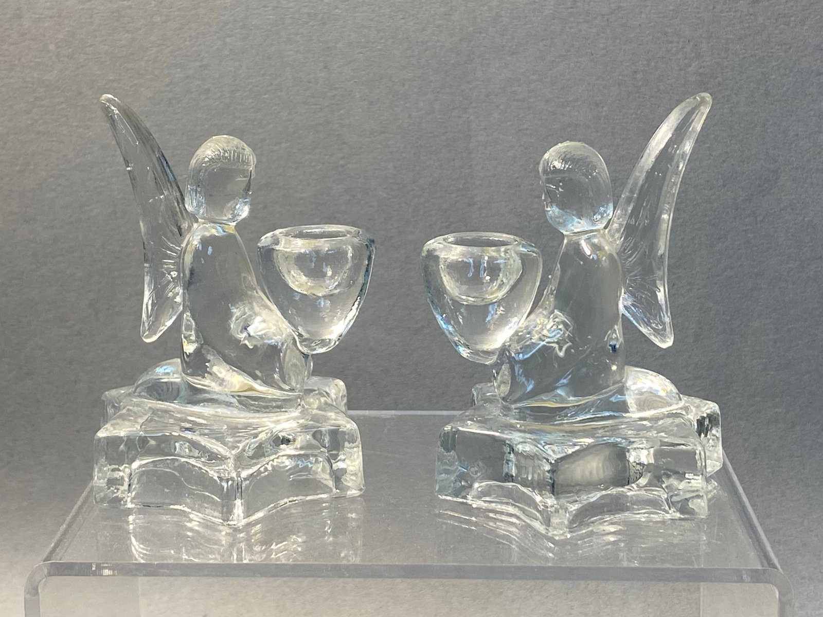 Pair of Bohemia Glass Angel Holders: A pair of Bohemia Glass clear molded glass candle holders manufactured in Czechoslovakia. Each figure depicts a kneeling angel holding a chalice-style cup while positioned on a six-pointed