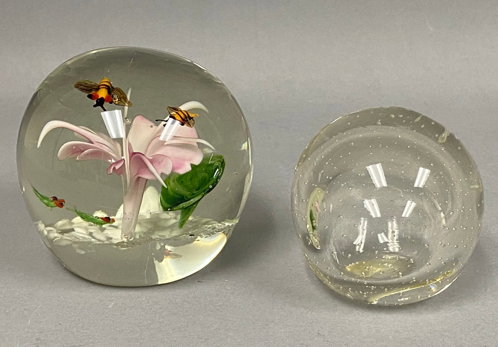 Two Hand Blown Art Glass Paperweights: This lot features two art glass paperweights showcasing different decorative techniques. The larger paperweight contains a detailed lampwork scene with a shaded pink flower, two yellow and black bees.