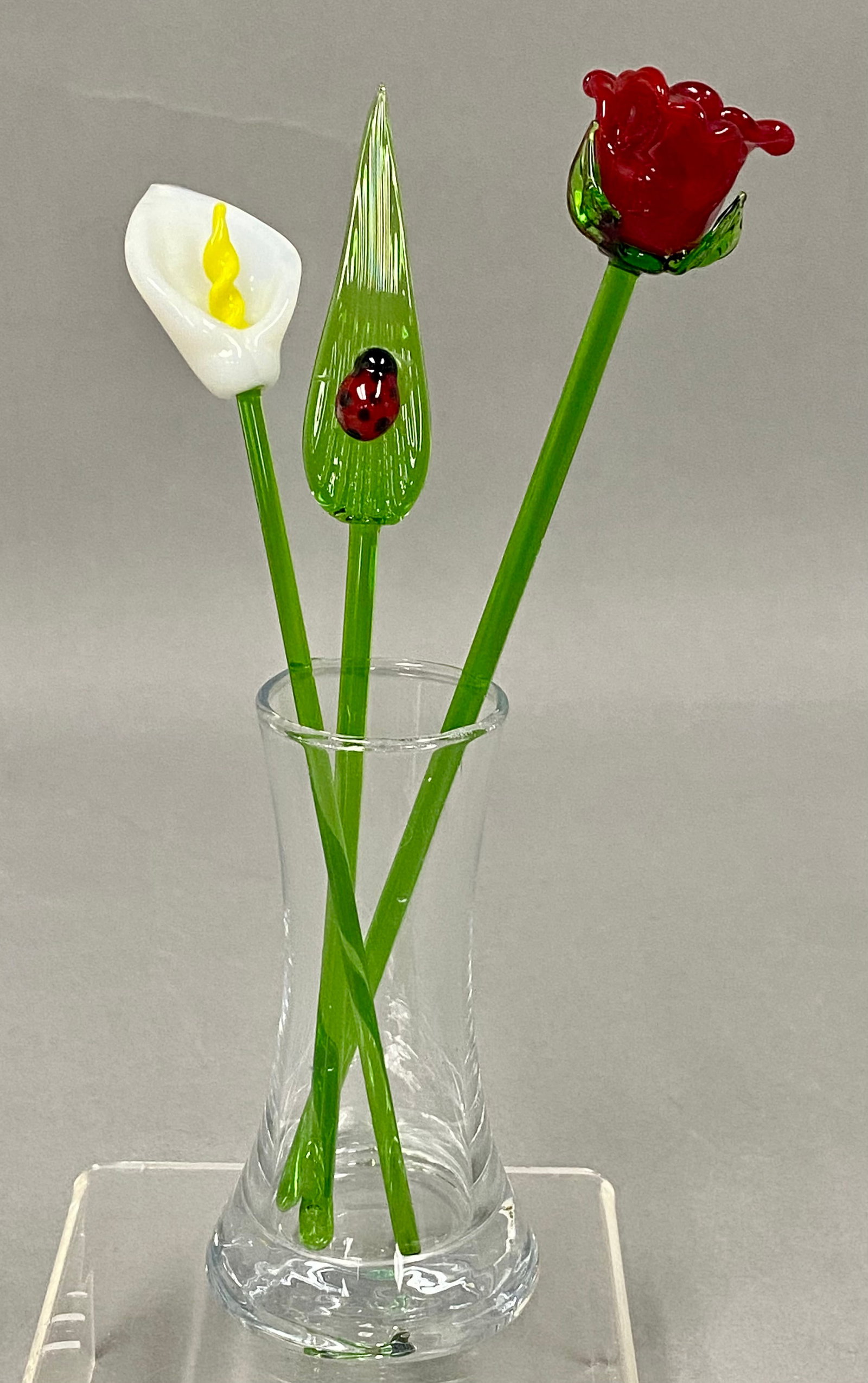 Red Co. Glass Flower Bouquet Set: This set consists of three flameworked glass stems including a white calla lily, a red rose, and a ribbed leaf with an applied red ladybug. Each piece is crafted from vibrant translucent and opaque