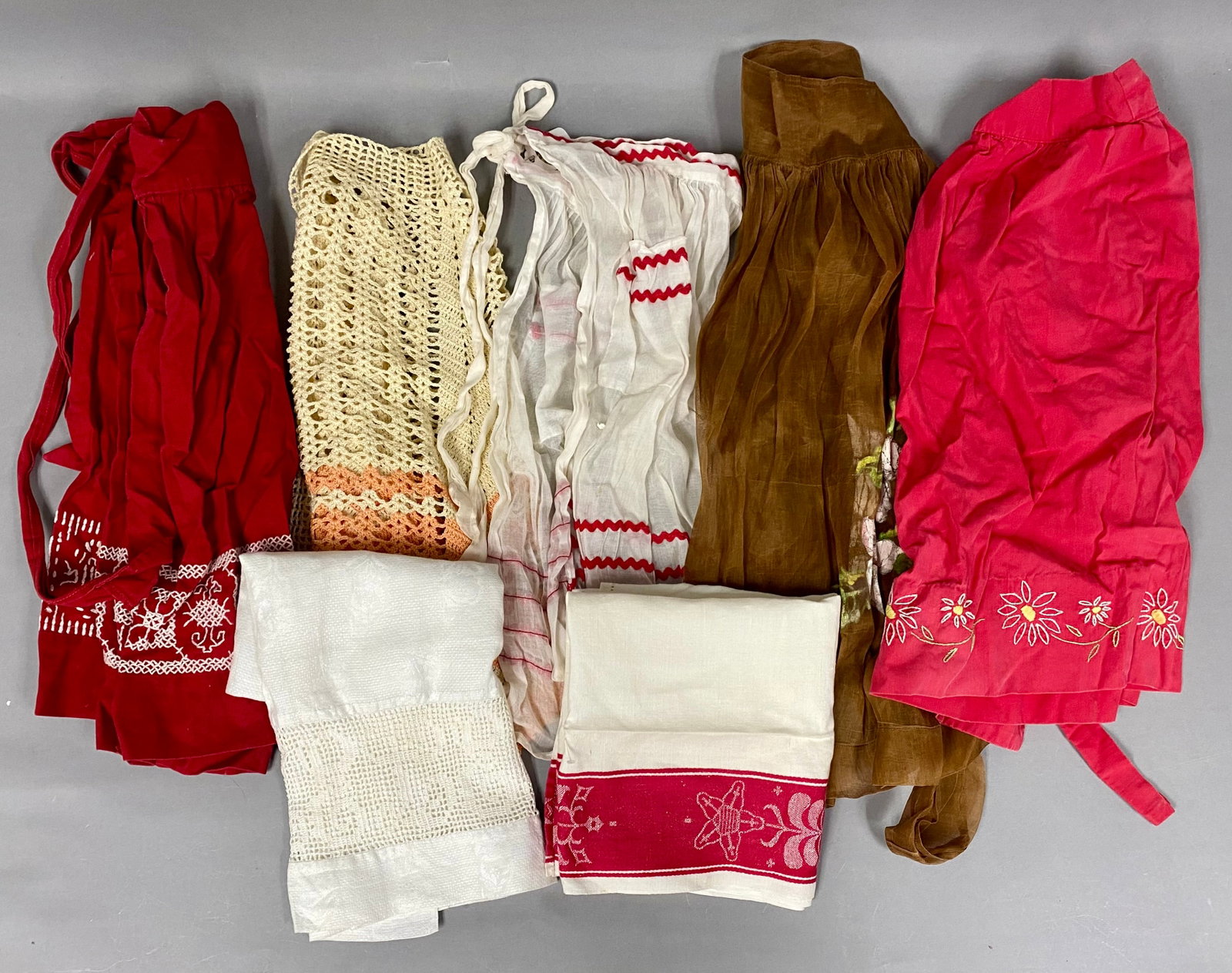 Collection of Seven Vintage Textiles: This collection includes five mid-century half-aprons and two linen hand towels showcasing various needlework techniques. The assortment features a hand-crocheted chevron apron, a hand-painted sheer