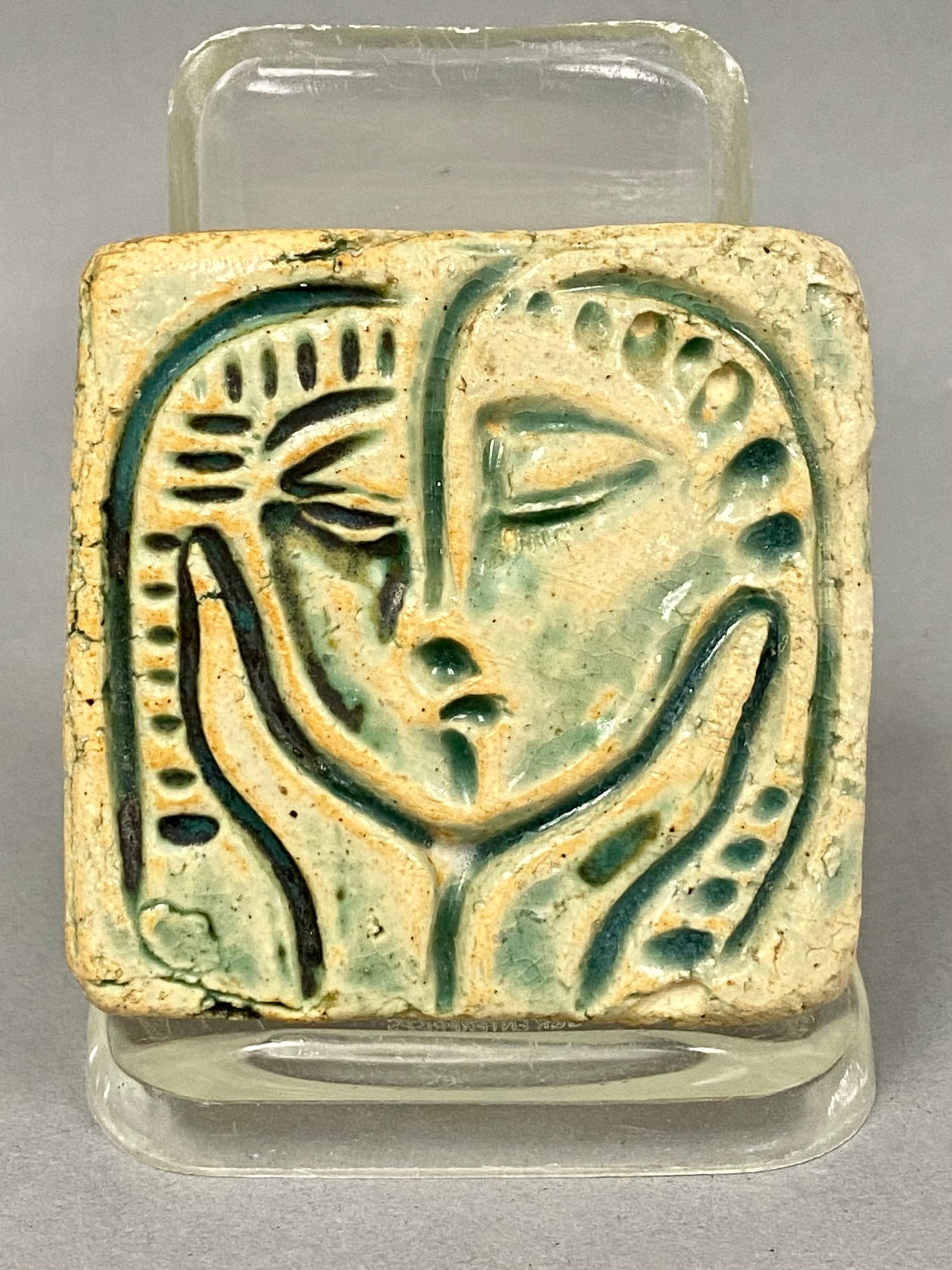 Pewabic Pottery Modernist Face Tile: This Pewabic Pottery earthenware tile features a relief-molded modernist design depicting overlapping faces cradled by a pair of hands. The surface is finished in a translucent green glaze with heavy
