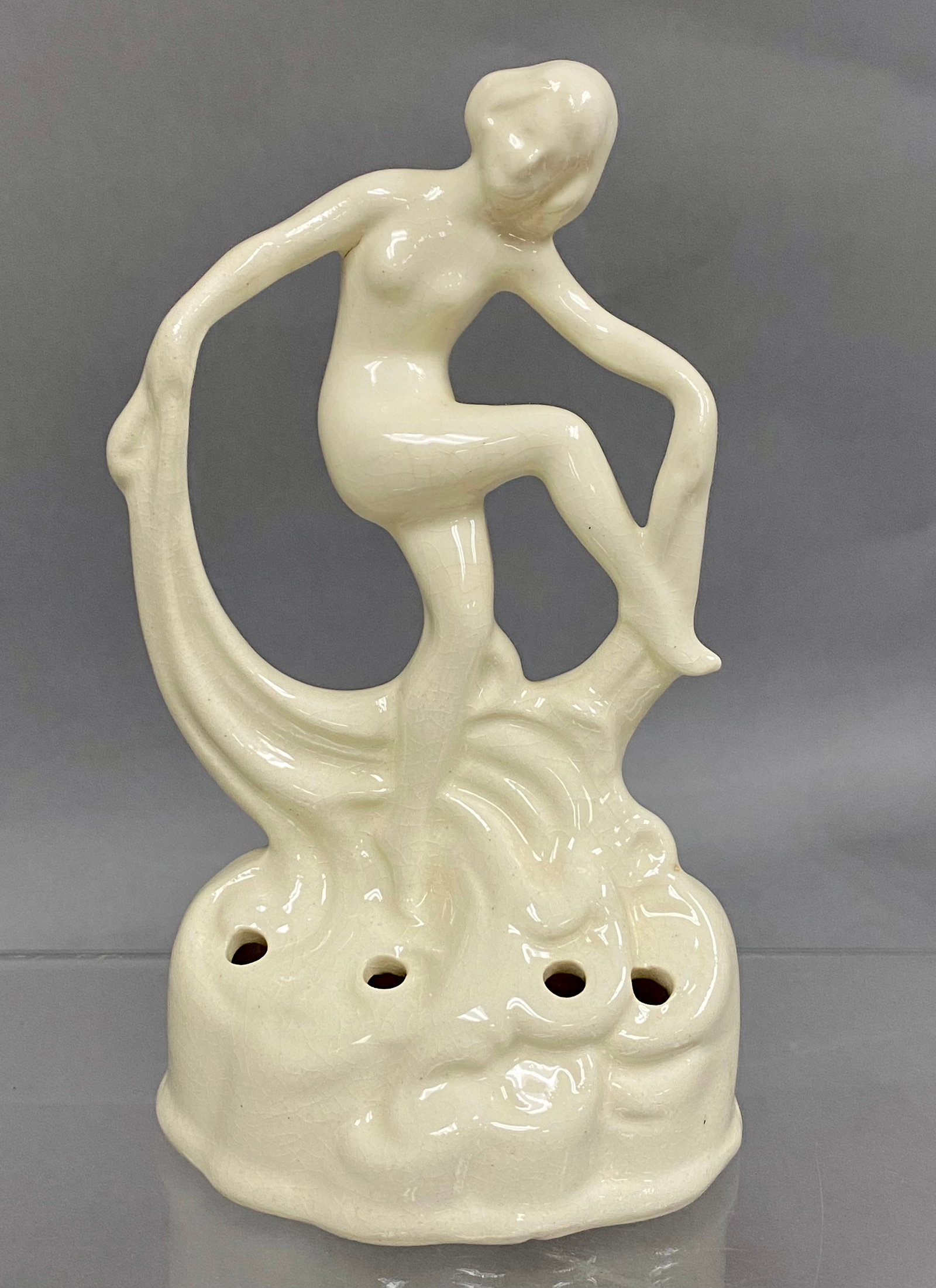 Art Deco Scarf Dancer Flower Frog: An Art Deco style cream glazed ceramic figurine depicting a stylized nude female dancer in a dynamic pose. The figure is modeled with a flowing scarf and stands upon a wave-inspired base perforated