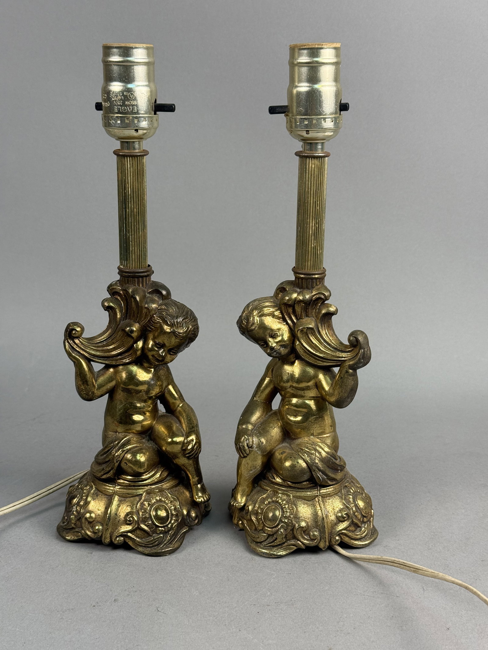 Gilt Cast Metal Cherub Table Lamps: A pair of table lamps featuring seated cherub figures in the Rococo Revival style. Each figure is cast in metal with a gilt finish and sits atop a highly ornate, scalloped base decorated with