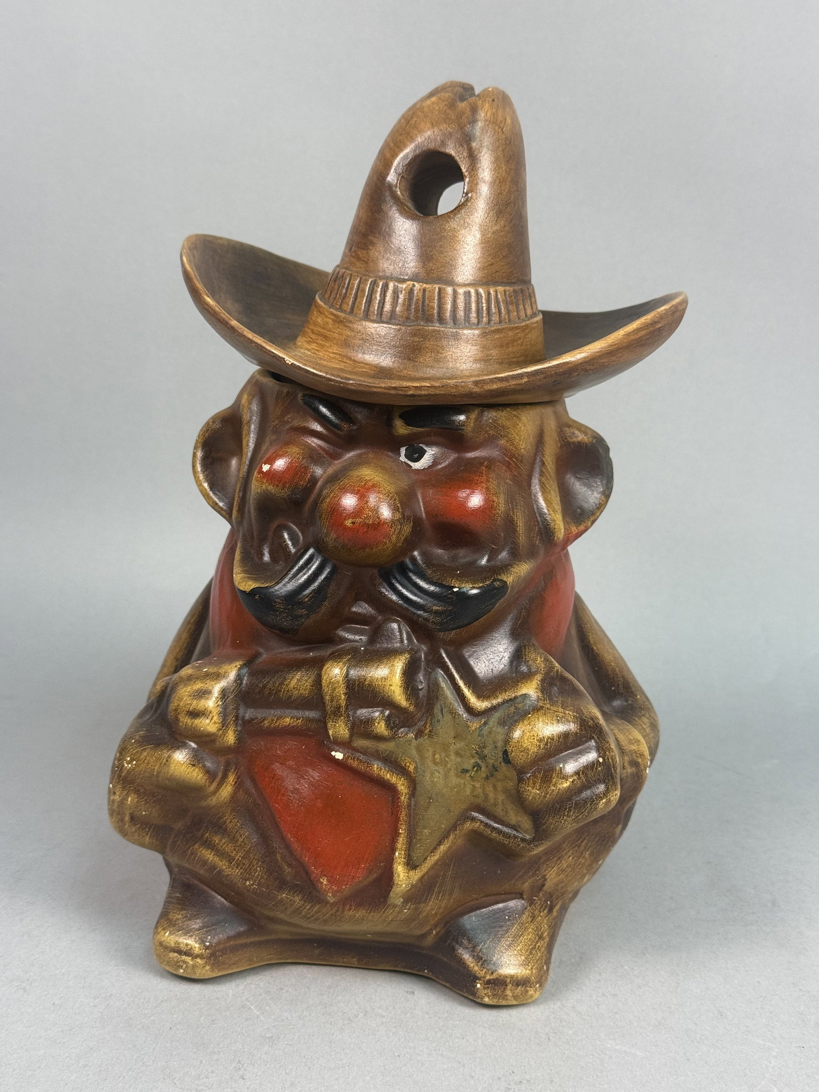 Ceramic Western Sheriff Cookie Jar: A vintage earthenware cookie jar depicting an anthropomorphic Western sheriff figure. The piece features a two-part construction, where the character's head and a large sombrero-style hat form the rem