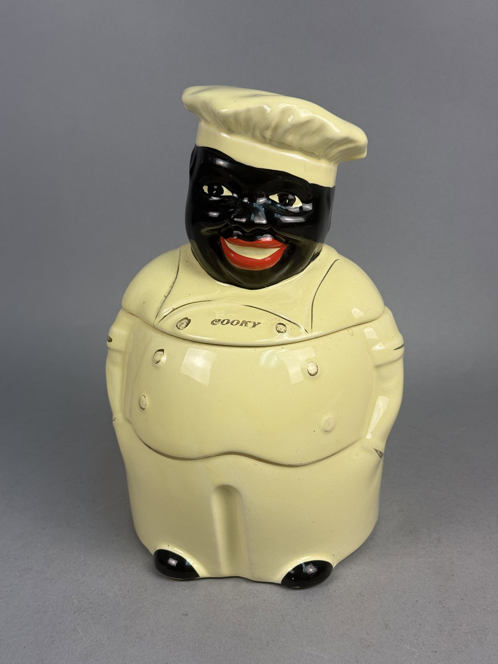 Pearl China Co Cooky Chef Ceramic Cookie Jar: A vintage two-piece glazed ceramic cookie jar in the form of a rotund chef, produced as a piece of Black Americana by the Pearl China Company. The figure is hand-painted with a pale yellow-cream