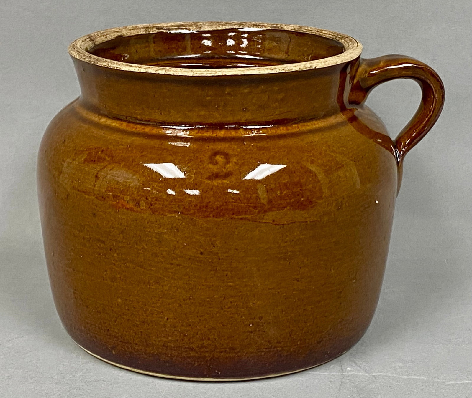 Brown Stoneware Single-Handled Bean Pot: A single-handled stoneware utility crock or bean pot finished in a glossy brown slip glaze. The piece features a bulbous body, a loop handle attached at the shoulder, and a recessed rim. The shoulder
