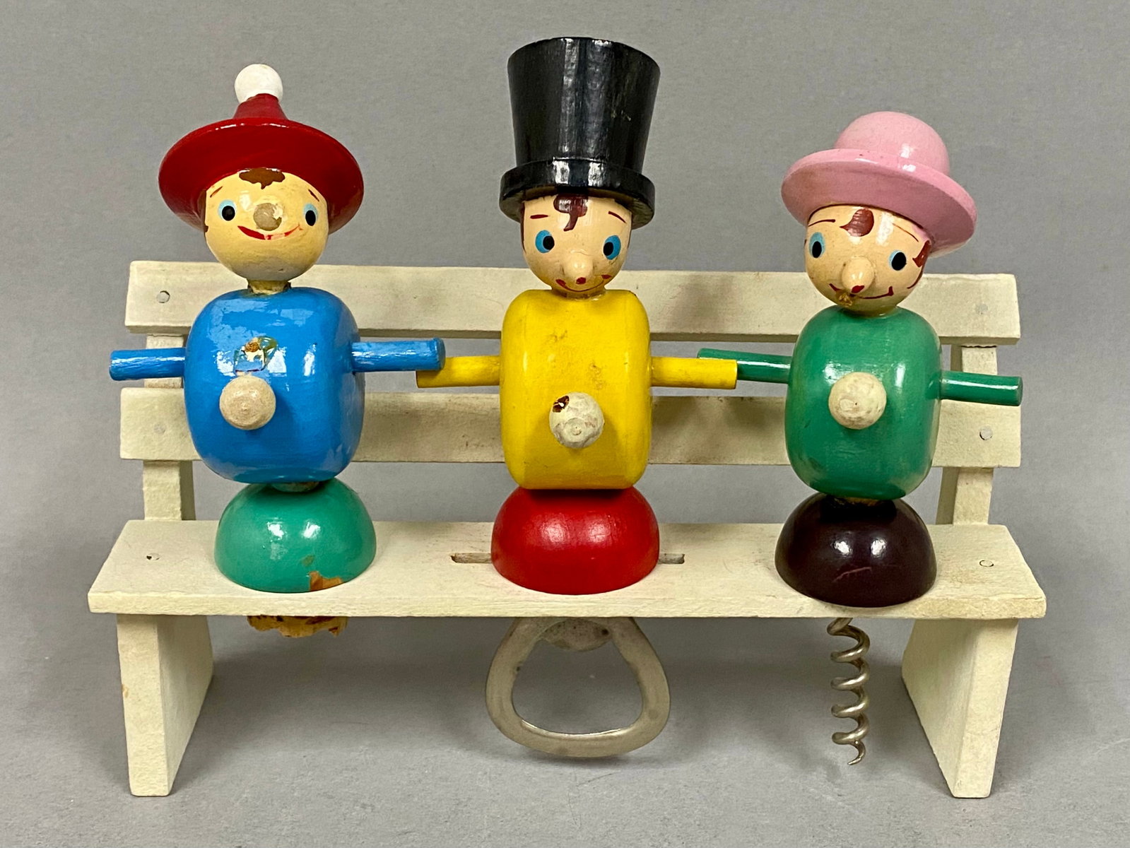 Mid-Century Wooden Character Bar Set: A mid-century modern wooden barware set featuring three anthropomorphic character figures serving as a bottle opener, corkscrew, and stopper. The tools are displayed on a white-painted wooden bench