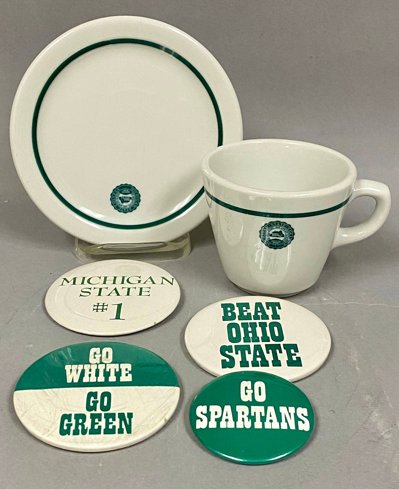 Shenango China Michigan State Memorabilia: This lot features a heavy vitrified ceramic cup and saucer set alongside a collection of collegiate pinback buttons. The white glazed china pieces are decorated with a forest green band and a