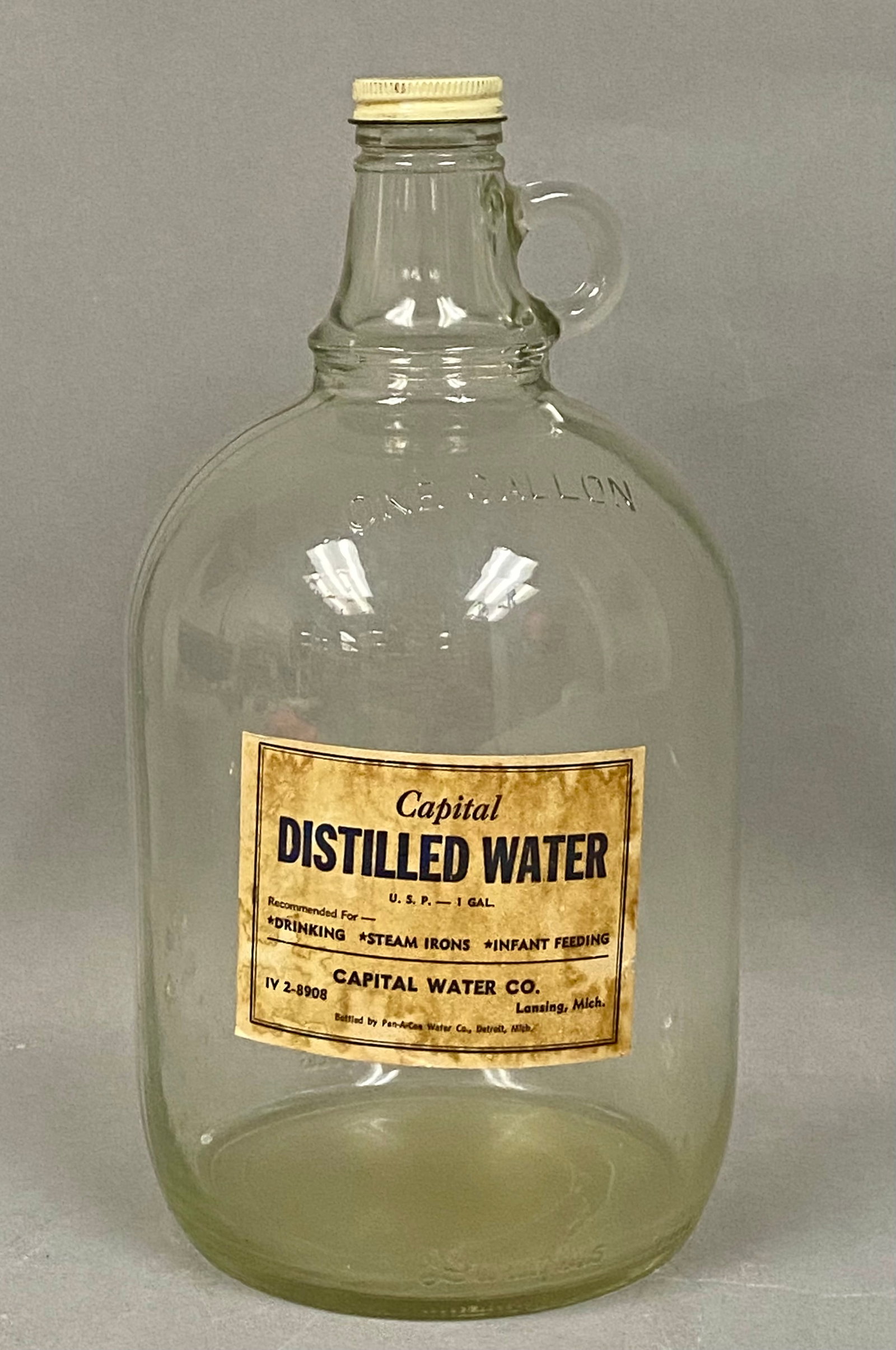 Vintage Capital Distilled Water Jug: A clear pressed glass one-gallon jug produced by the Owens-Illinois Glass Company featuring an original paper label for Capital Distilled Water of Lansing, Michigan. The body is embossed with the