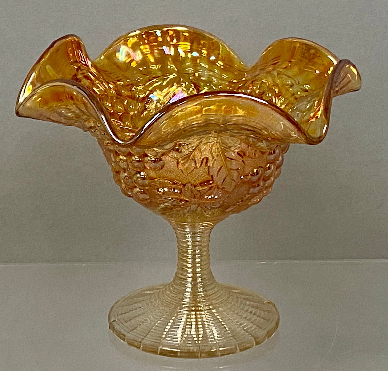 Marigold Carnival Glass Ruffled Compote: An American marigold carnival glass footed compote featuring a ruffled rim and the Grape and Cable pattern. The exterior bowl is decorated with high-relief grape clusters and leaves joined by a cable