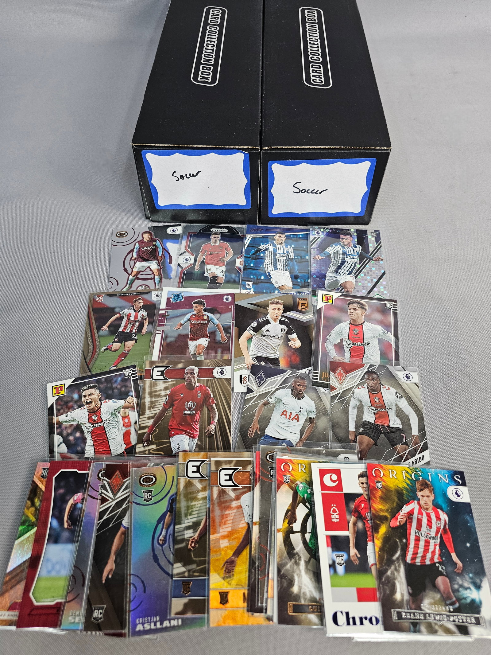 Assorted Soccer Trading Card Collection: An assortment of soccer trading cards primarily produced by Panini, featuring various modern sets housed in two black cardboard boxes. Featured players include Harry Kane, Heung-Min Son, Jack Grealish