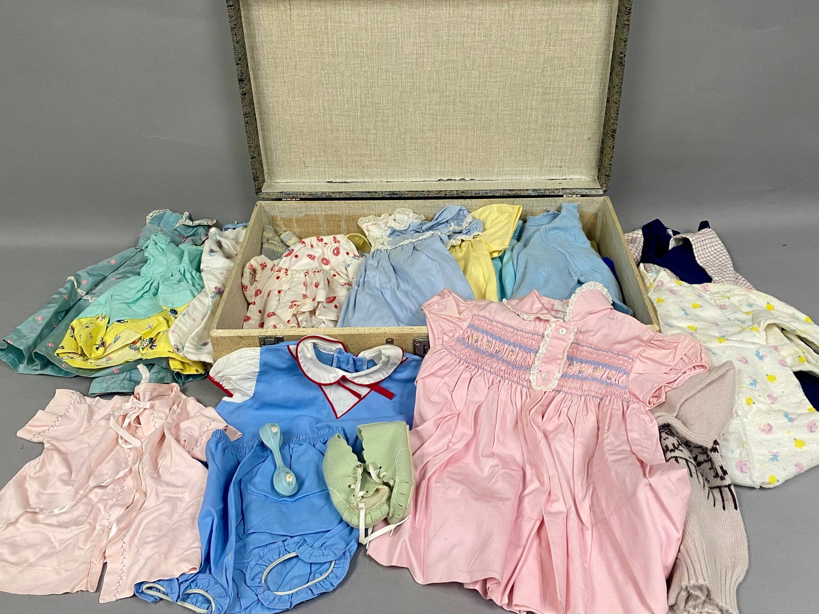 Vintage Doll and Infant Clothing: An assortment of vintage infant and doll-sized garments and accessories housed in a mid-century hard-sided suitcase. The collection features a pink satin-finish top bearing a cloth tag that reads Kitt