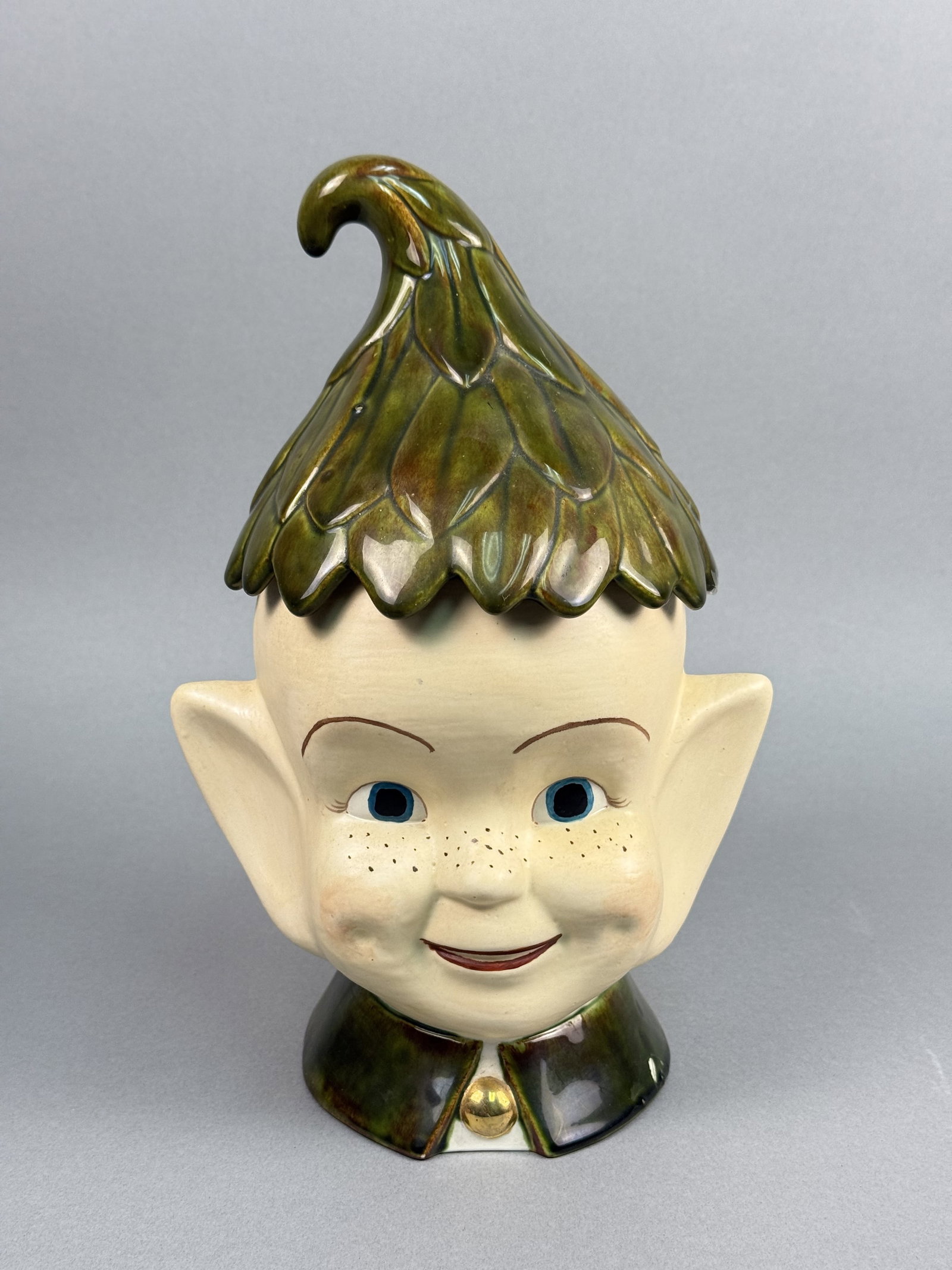 Gerry Ceramic Pixie Cookie Jar: A mid-century two-piece glazed ceramic cookie jar designed in the form of a whimsical pixie or elf head. The vessel features a removable lid shaped as a pointed green cap with textured leaf-like