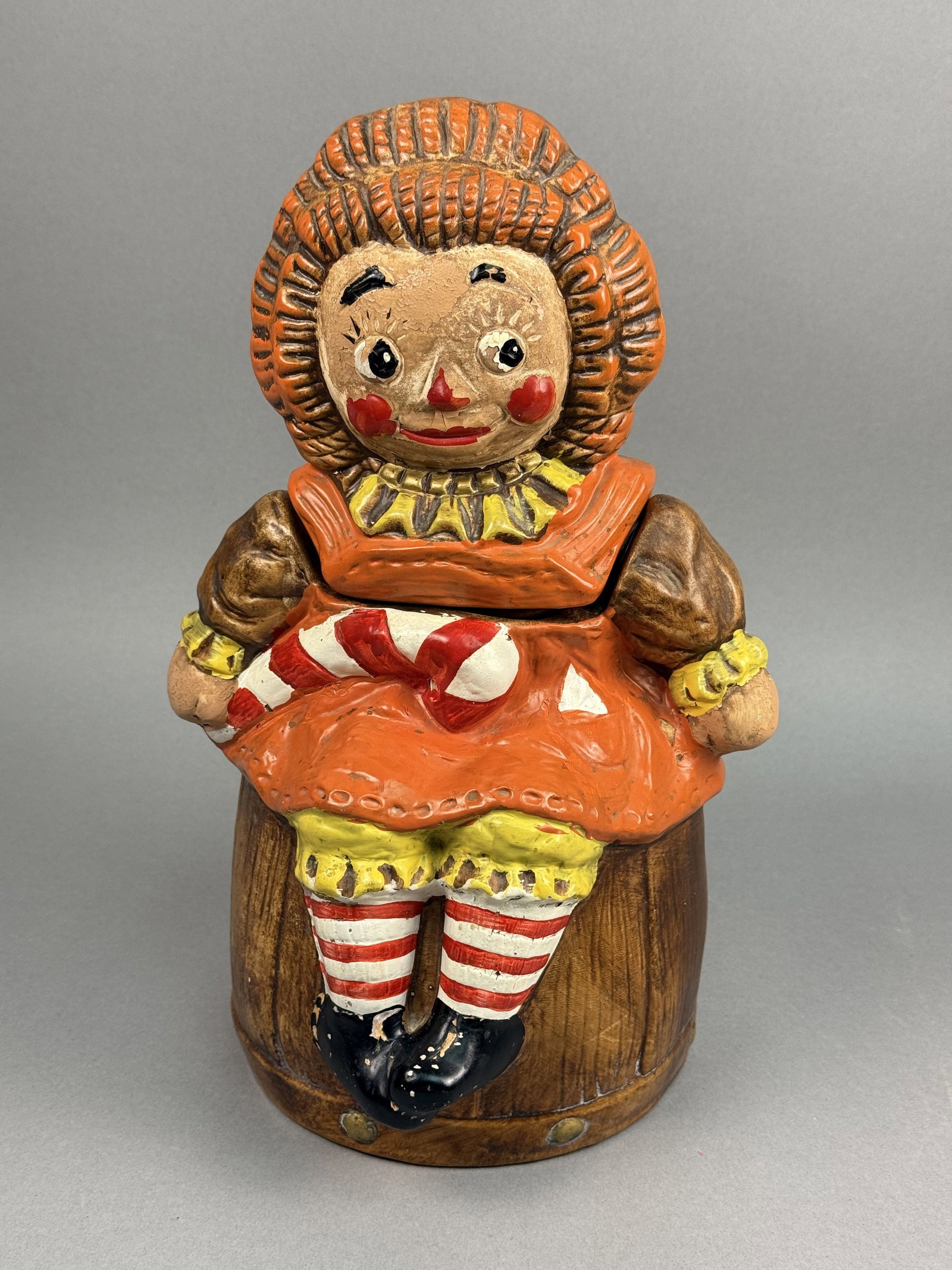 Hand-Painted Ceramic Raggedy Ann Cookie Jar: A vintage ceramic cookie jar featuring the character Raggedy Ann seated on a textured barrel. The interior of the piece is finished in a clear glaze. The exterior is decorated with cold-painted
