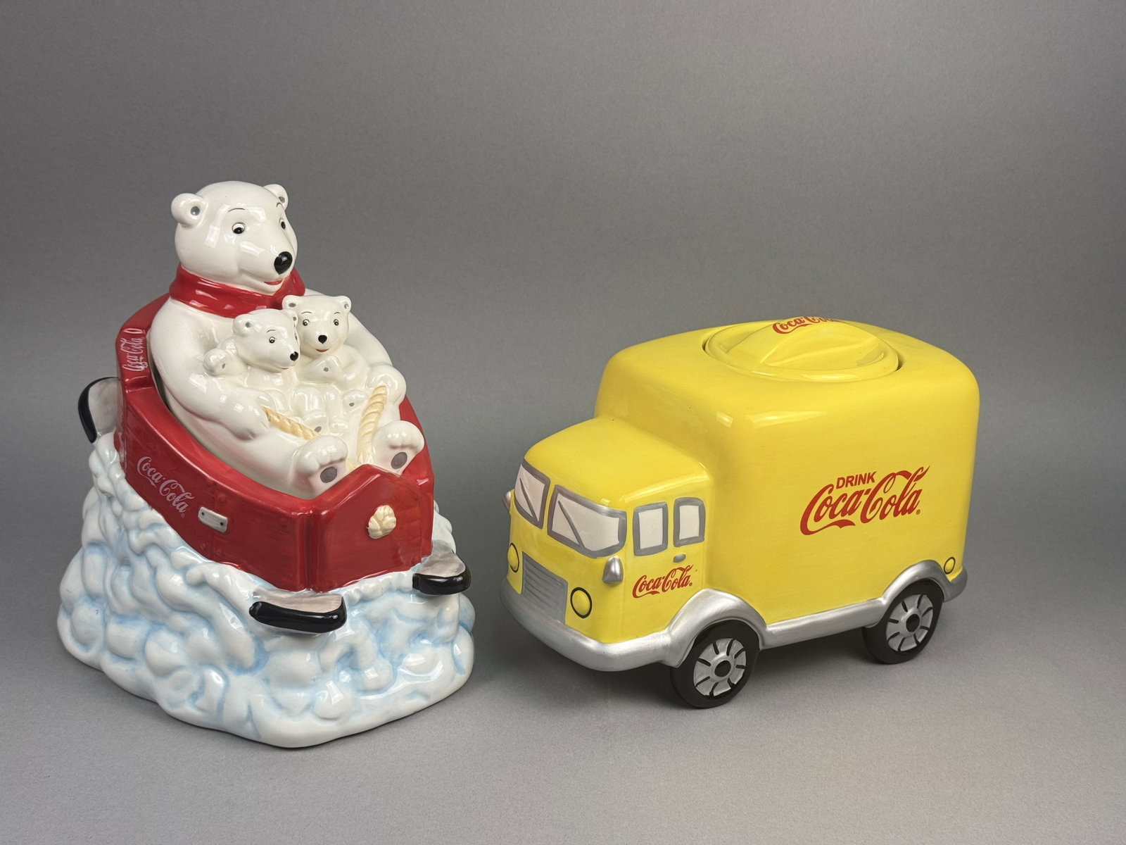 Gibson Coca-Cola Ceramic Cookie Jars: Two licensed Coca-Cola earthenware cookie jars with their original retail packaging. The first jar depicts an adult polar bear wearing a red scarf, accompanied by two cubs in a red sled mounted on a w