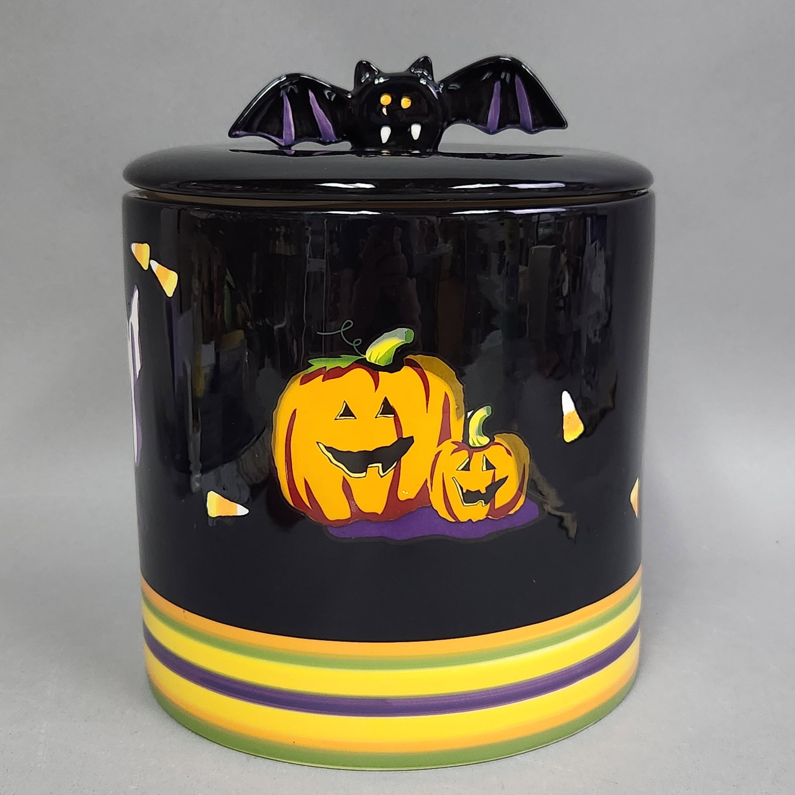 Halloween BOOlevard Bat Handle Cookie Jar: This Chinese-made glazed ceramic cookie jar from the Halloween BOOlevard line features a cylindrical form with a glossy black ground. The inset lid is topped with a figural bat handle detailed with pu