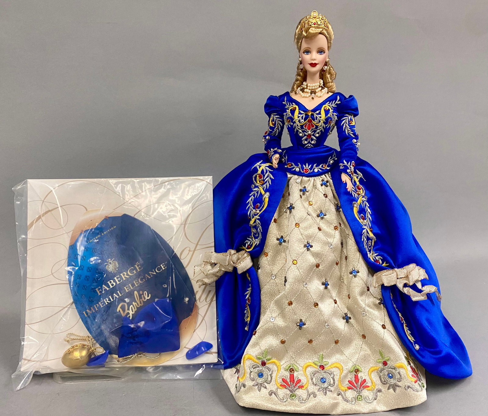 Mattel Faberge Imperial Elegance Vinyl Barbie Doll: This Mattel limited edition porcelain doll is the Faberge Imperial Elegance Barbie, released in 1998. The figure is dressed in an ornate royal blue velvet-style gown featuring elaborate gold-tone embr