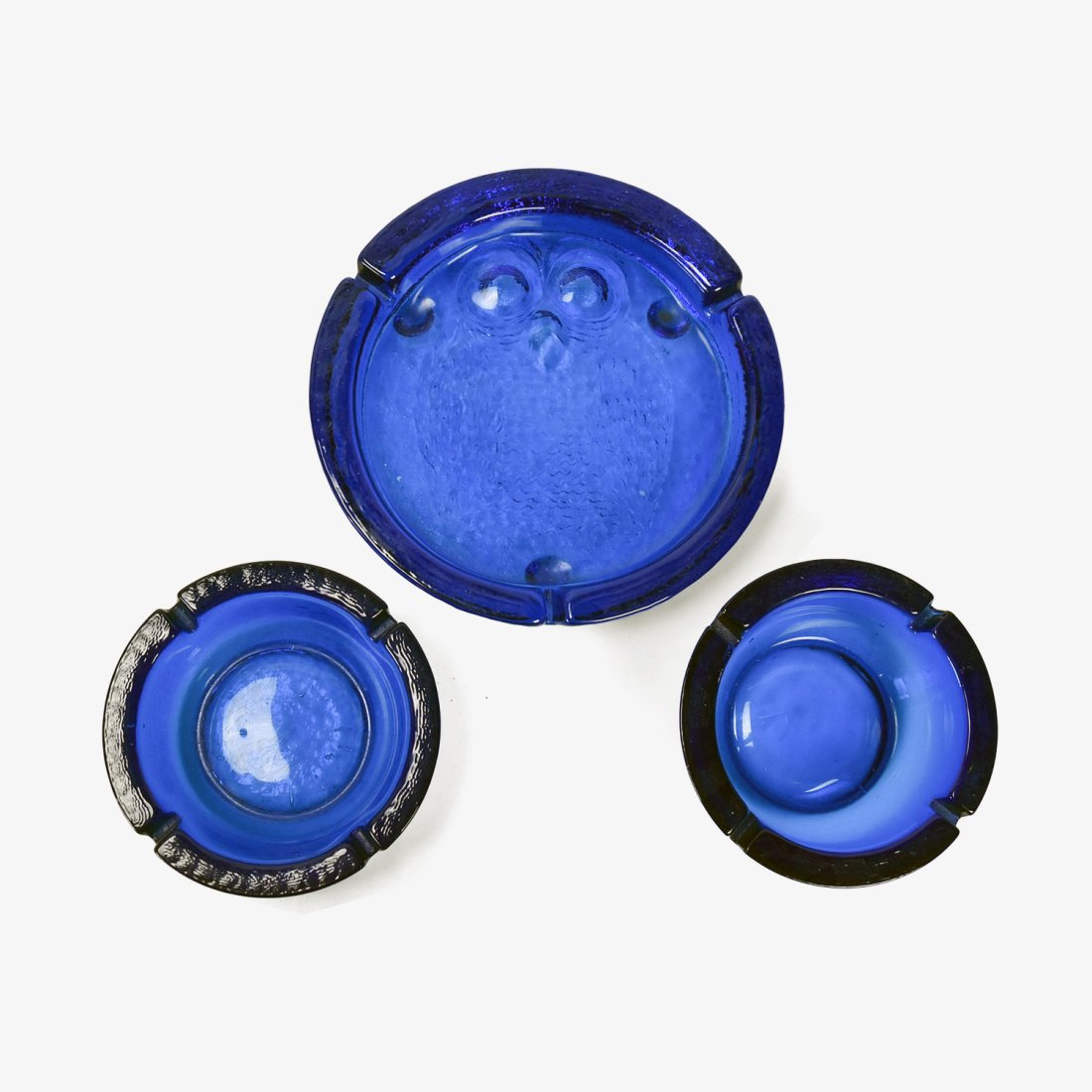 Lot of Three Blenko Cobalt Blue Ashtrays: Featured are three Blenko cobalt blue ashtrays, 1 larger ashtray, and two smaller identical ashtrays. The larger of the ash trays features an owl design with the top inside area raised for the owls ey