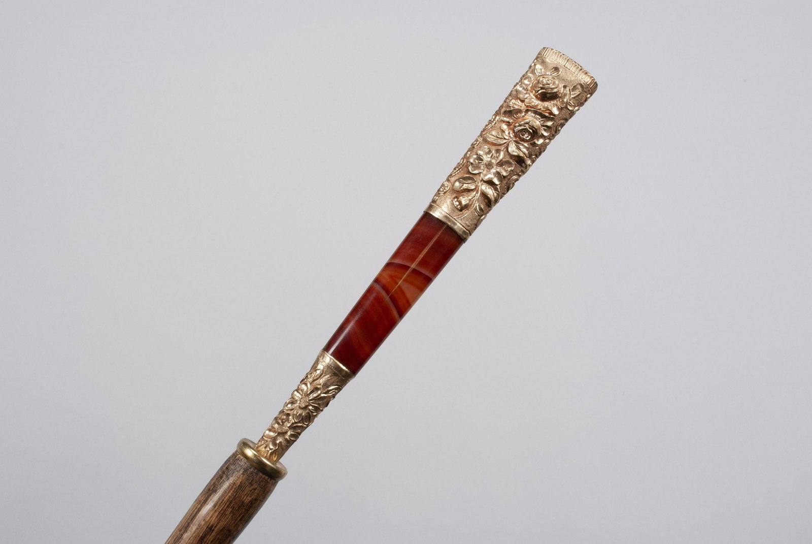 A 14K MOUNTED WALKING STICK/CANE: A 14K MOUNTED WALKING STICK/CANE With floral and rosette decorated handle marked 14k with "L" inside triangle, bookending a section of banded red agate, and mounted to wooden shaft with brass ferrule.