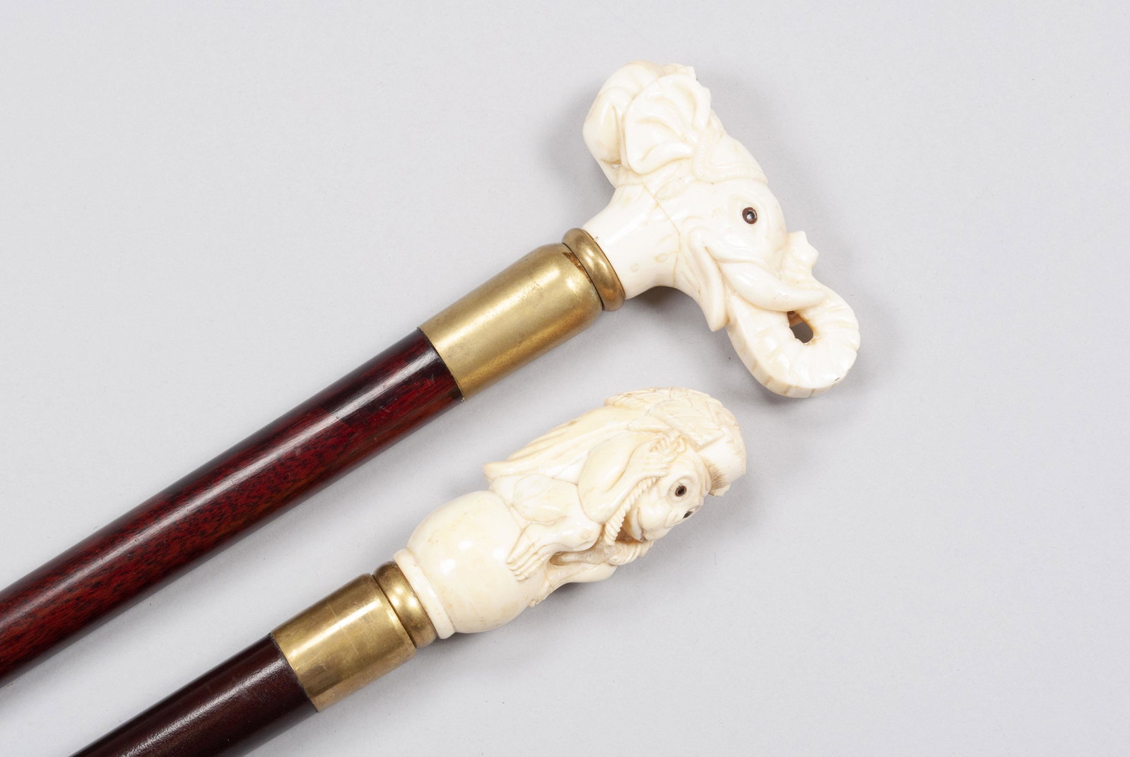 PAIRING OF CARVED WALKING STICKS/CANES: PAIRING OF CARVED WALKING STICKS/CANES Both with carved and segmented finials with glass eyes atop brass fittings on lacquered, dark wooden shafts: one of a monkey with a basket held over his head (lo