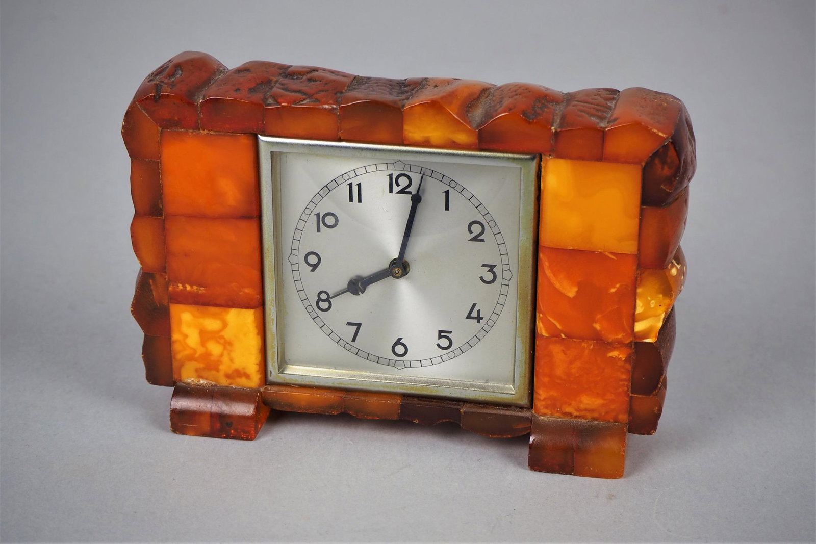 Art deco amber clock, Staatliche Bernstein-Manufaktur, Königsberg, around 1930.: Art deco amber clock, Staatliche Bernstein-Manufaktur, Königsberg, around 1930.Small square table clock to wind up in a rectangular wooden plate, covered all around with pieces of amber with a mo