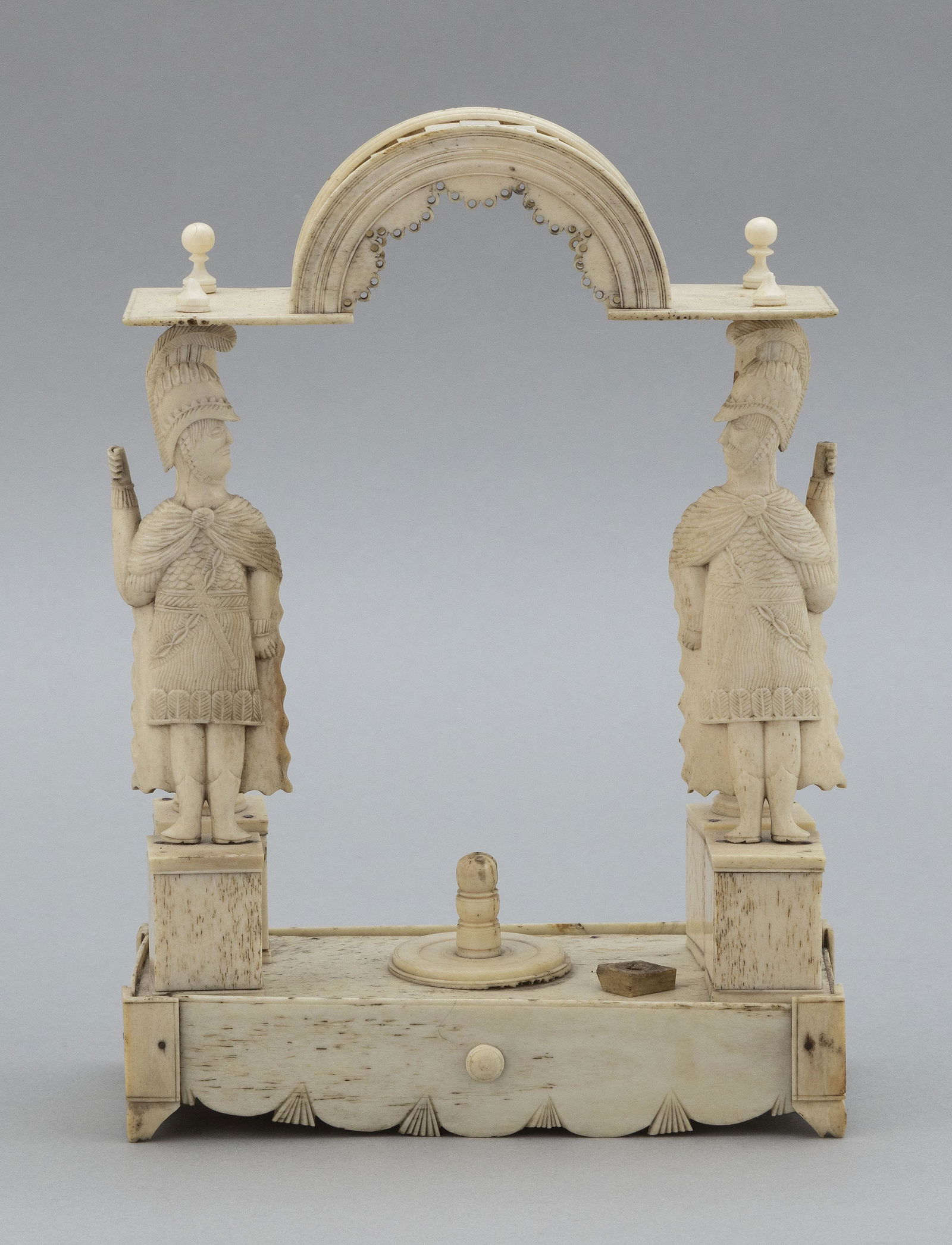 NAPOLEONIC PRISONER-OF-WAR CARVED BONE STAND: NAPOLEONIC PRISONER-OF-WAR CARVED BONE STANDFrench/English, 19th CenturyRectangular footed base with two helmeted warriors on plinths and two tapered columns supporting an arched roof with finials at