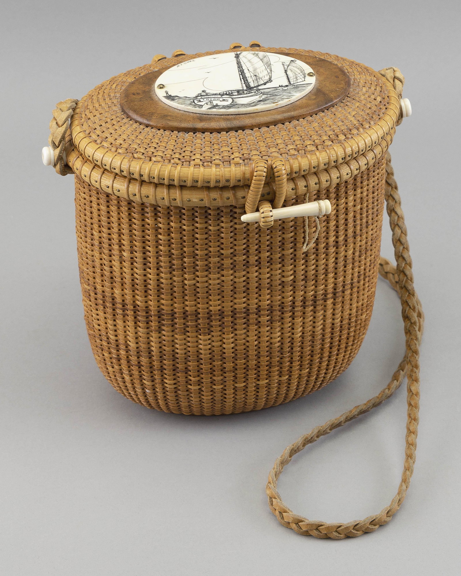 NANTUCKET BASKET PURSE BY GERALD L. BROWN Nantucket, Massachusetts, Contemporary Height 8.25”.: NANTUCKET BASKET PURSE BY GERALD L. BROWN Nantucket, Massachusetts, Contemporary Oval swallow’s-nest basket with woven leather strap. Scrimshaw-style engraved plaque on lid depicts a catboat, si