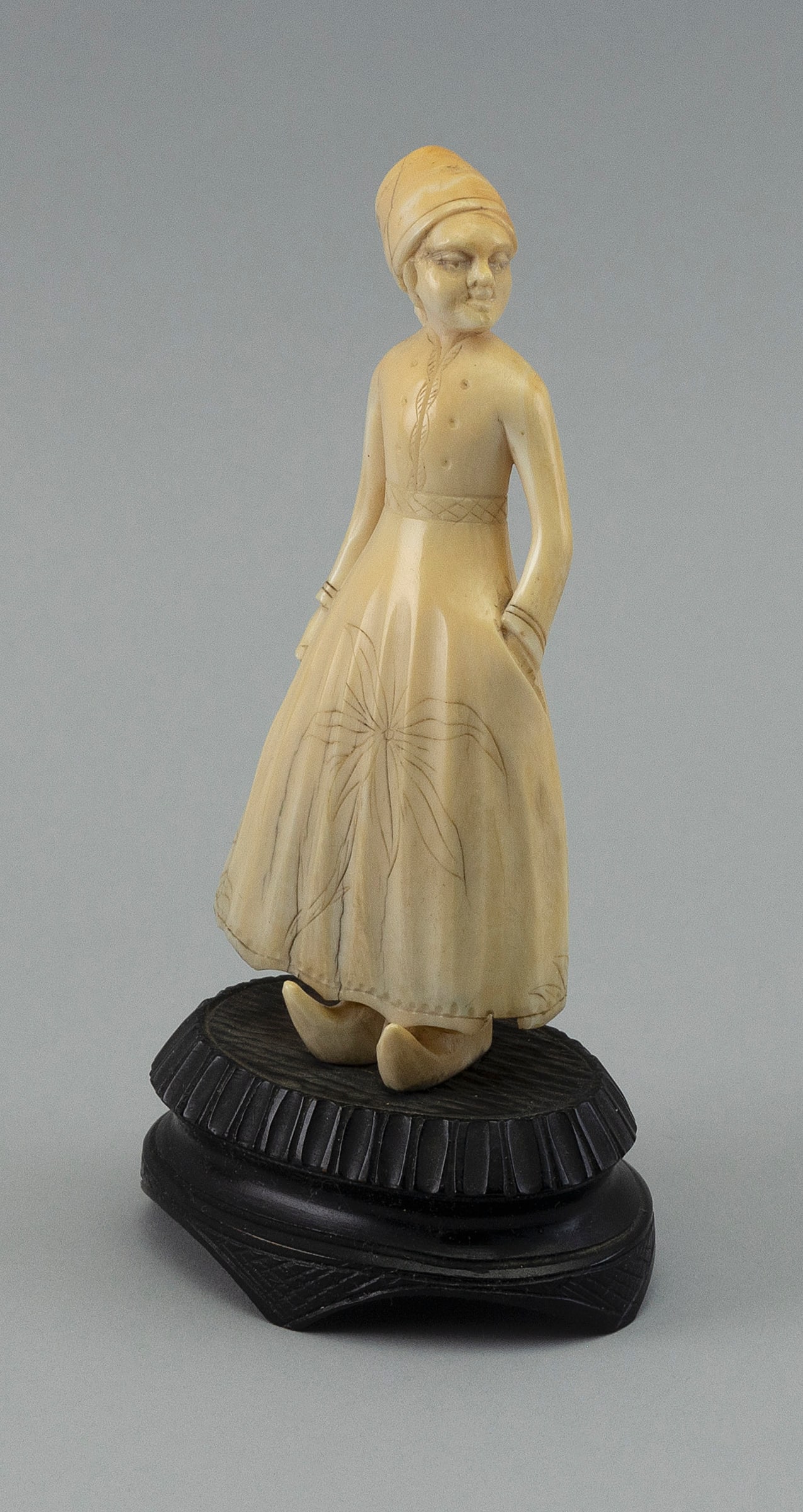 CARVED WHALE IVORY FIGURE OF A DUTCH GIRL 19th Century Total height 6.25".: CARVED WHALE IVORY FIGURE OF A DUTCH GIRL 19th Century Carved from a whale's tooth. Girl wearing a bonnet, a dress with an engraved floral design on skirt, and traditional clogs. Wooden stem and eboni