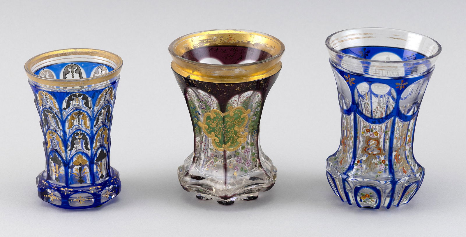 THREE BOHEMIAN BIEDERMEIER CUT OVERLAY GLASS BEAKERS Mid- to Late 19th Century Heights 4.5", 5" and: THREE BOHEMIAN BIEDERMEIER CUT OVERLAY GLASS BEAKERSMid- to Late 19th CenturyTwo cobalt blue to clear and one lavender to clear, all with enameled and gilt decoration. One cobalt blue also with etched