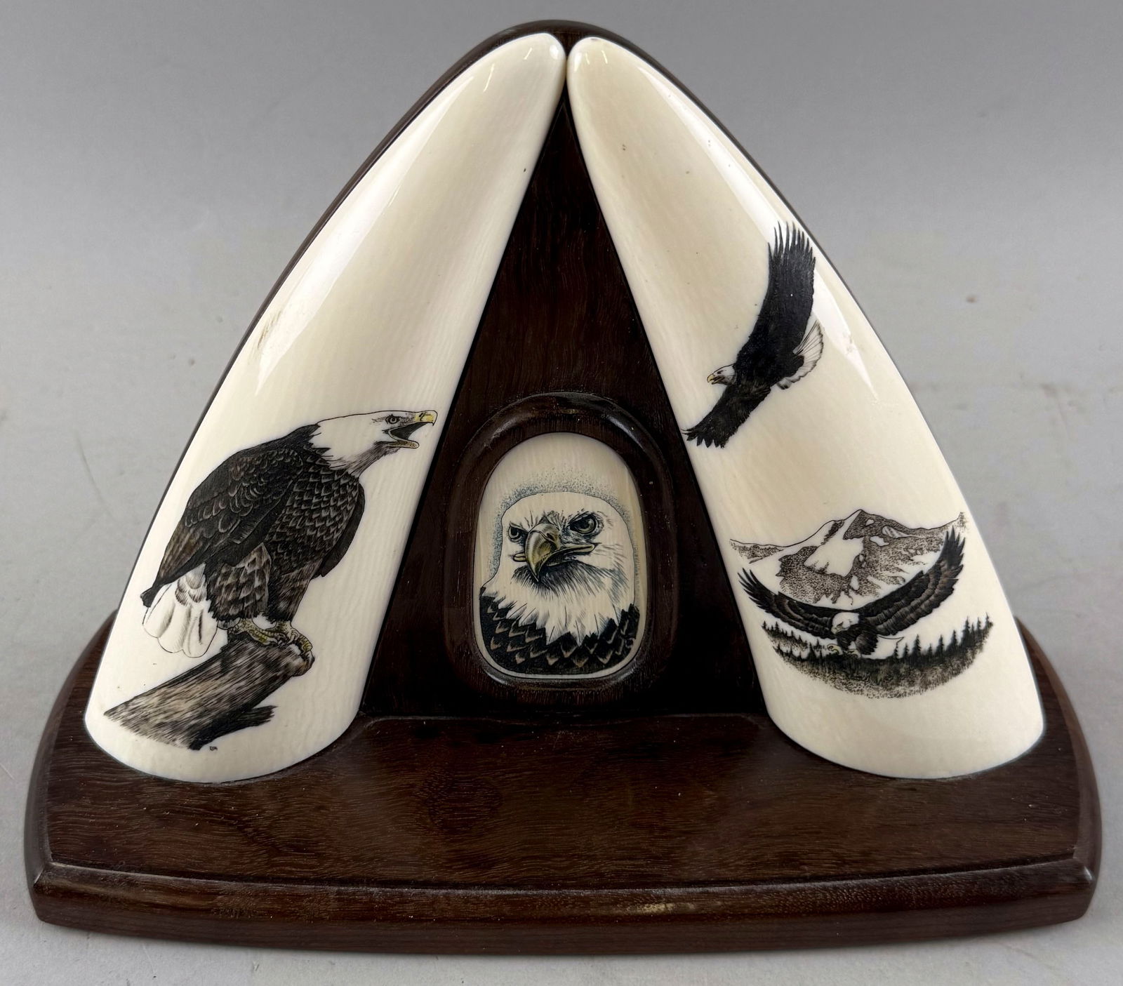 PAIR OF MOUNTED AND ENGRAVED SIMULATED WHALE'S TEETH 20th Century Height 7.5". Length 9.5". Depth: PAIR OF MOUNTED AND ENGRAVED SIMULATED WHALE'S TEETH20th CenturyTeeth polychrome engraved with eagles and mounted on a hardwood base flanking a simulated ivory medallion, also polychrome engraved with