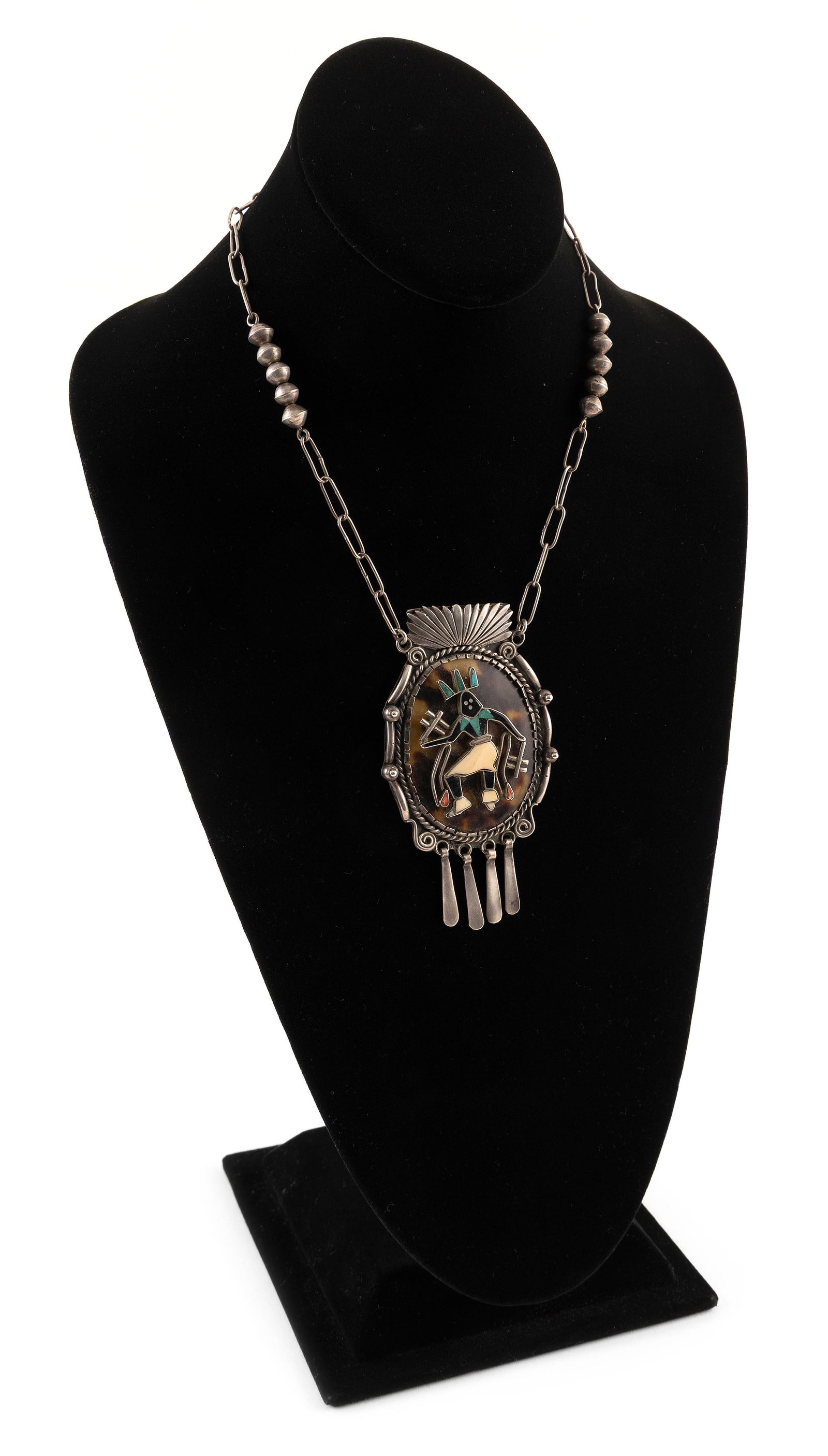 ANDREW DEWA ZUNI STERLING SILVER NECKLACE 20th Century Length 11".: ANDREW DEWA ZUNI STERLING SILVER NECKLACE20th CenturySilver-framed drop pendant with four dangles and a Kachina dancer comprised of turquoise, jet, coral and shell. Silver open-link chain with bead sp