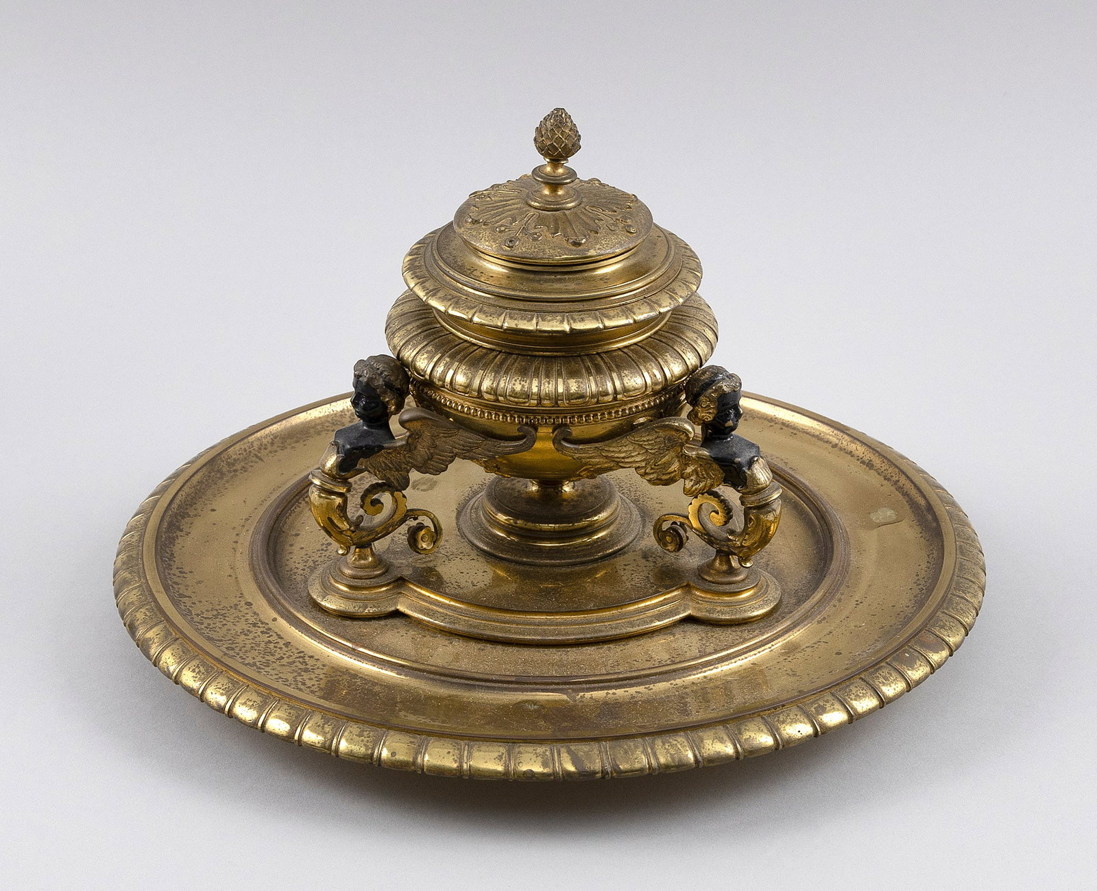 GILT METAL INKWELL Late 19th Century Height 7". Diameter 10".: GILT METAL INKWELL Late 19th Century Formed as a Grecian urn with three winged cherub supports and a gadrooned base. Lid with an acorn knop. Height 7". Diameter 10".