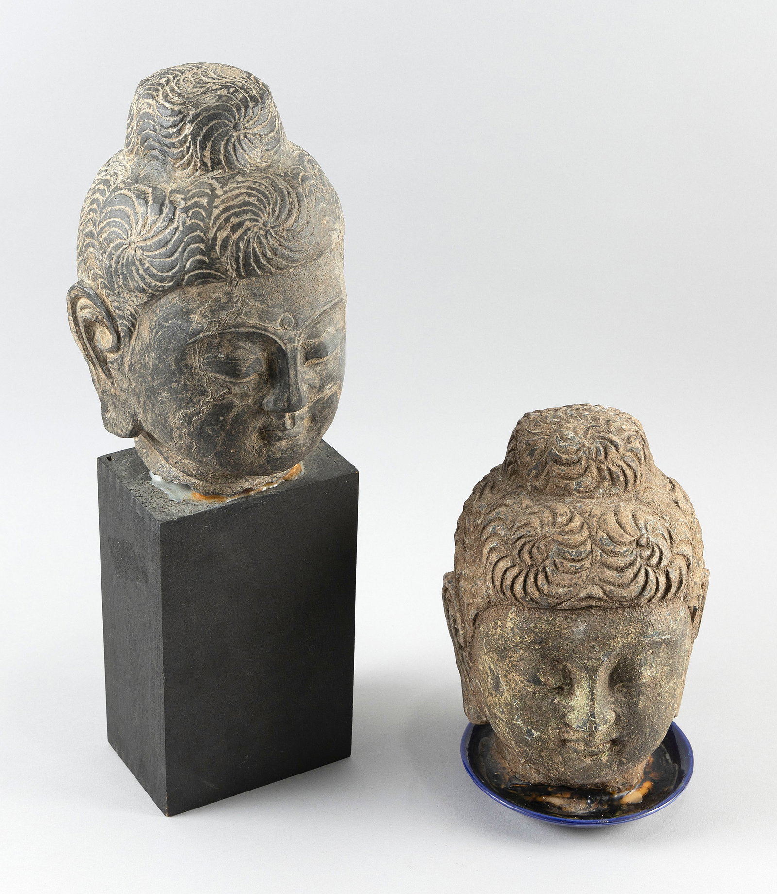 TWO ARCHAIC GANDHARAN-STYLE CARVED SANDSTONE BUDDHA HEADS: TWO ARCHAIC GANDHARAN-STYLE CARVED SANDSTONE BUDDHA HEADS 1) Mounted on a wood plinth. Head height 10". Total height 18.5". 2) Affixed to a blue saucer-type base. Total height 9".