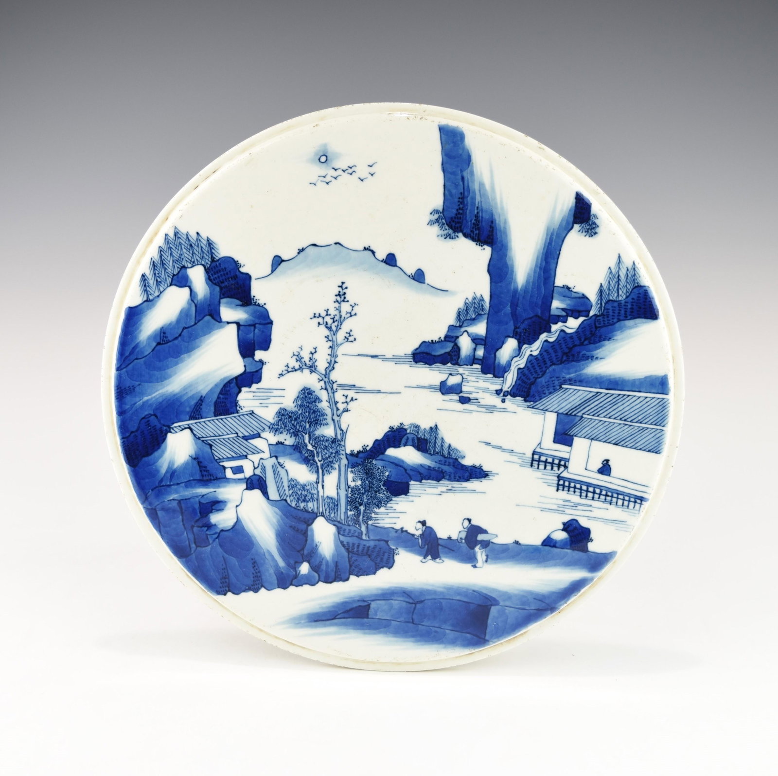 A 19TH CENTURY CHINESE BLUE AND WHITE DISC TILE: Decorated with mountainous landscape with cottages by riverside, birds flying in the sky under sun, and two travelers on the riverside road. Condition: This tile disc has a couple of firing cracks as 