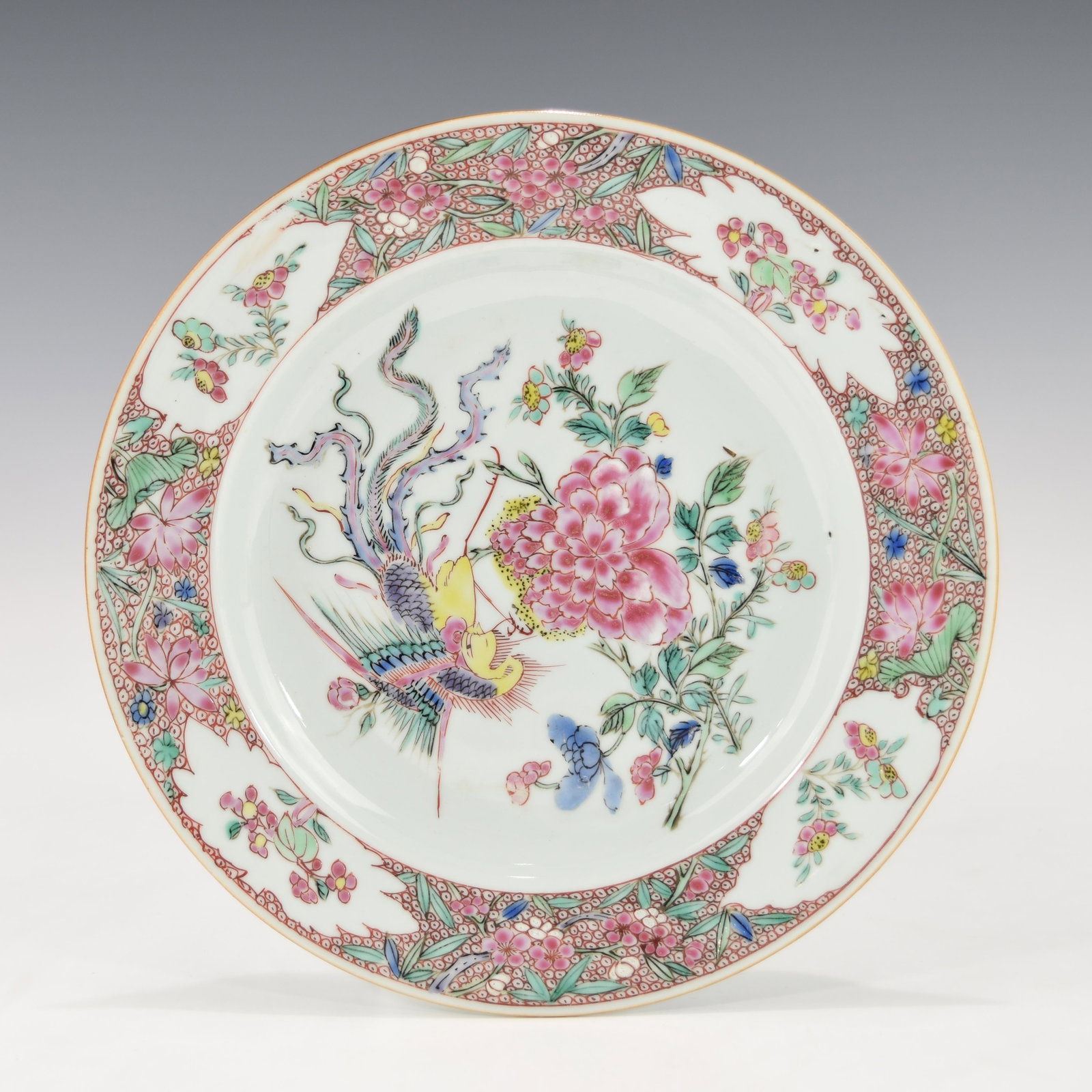 A YONGZHENG PERIOD (1722-1735) FAMILLE ROSE PLATE: Painted in soft vibrant famille rose enamel, depicting a phoenix flying around flowering bushes in the center, a band of alternating leaf-shaped cartouches, lily and prunus flowers along the outer rim