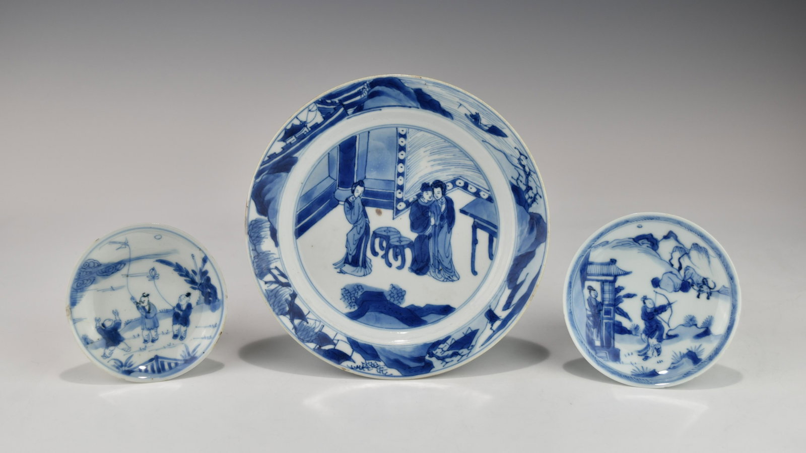 THREE CHINESE KANGXI PERIOD BLUE AND WHITE PORCELAIN DISHES: Decorated with various figural scenes at the front. The larger one is in very good condition with minor frittings or fleabites along the top rim, and a six-character Chenghua mark at the base. Both of