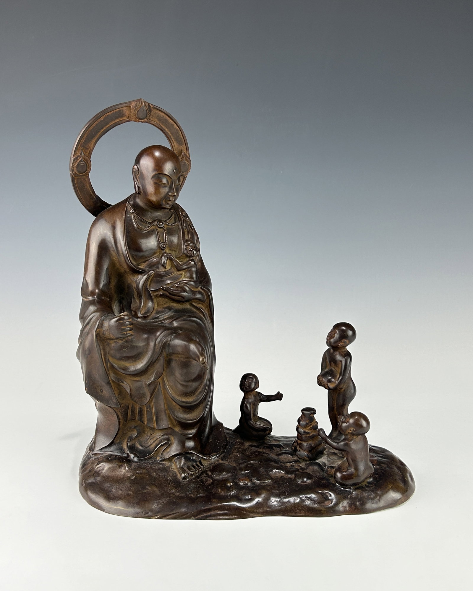 ANTIQUE JAPANESE BRONZE BUDDHA WITH CHILDREN: 19TH/20TH century, featuring a serene figure of Buddha seated with a group of children gathered around him. The Buddha, depicted with a calm and compassionate expression, is draped in flowing robes an