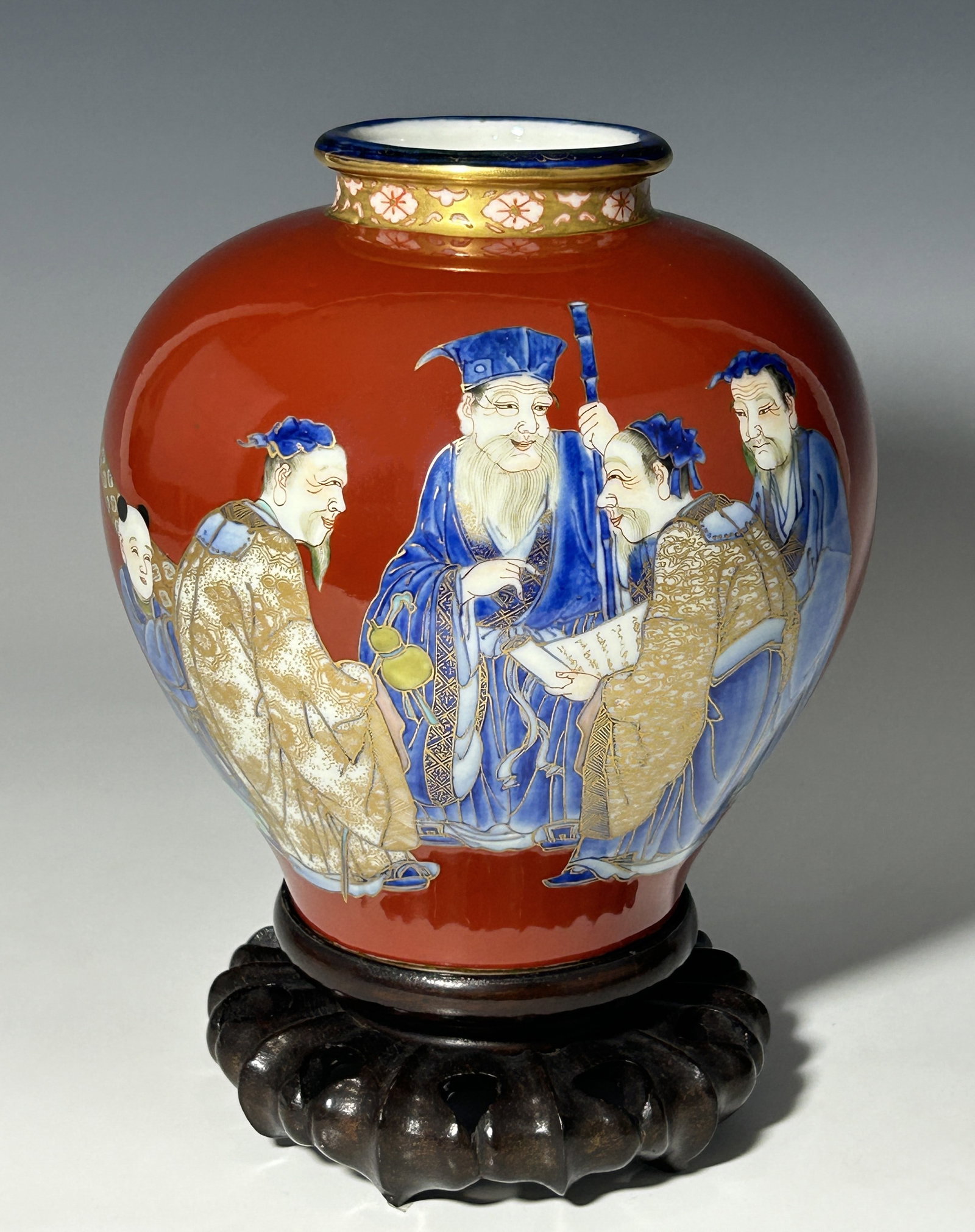 ANTIQUE JAPANESE MEIJI PERIOD FUKAGAWA PORCELAIN JAR PORTRAYING SCHOLAR GATHERING: This antique Japanese porcelain jar captures a dynamic scene of scholars in an engaged gathering, accompanied by a servant standing attentively nearby. Set against a deep red background, the figures a
