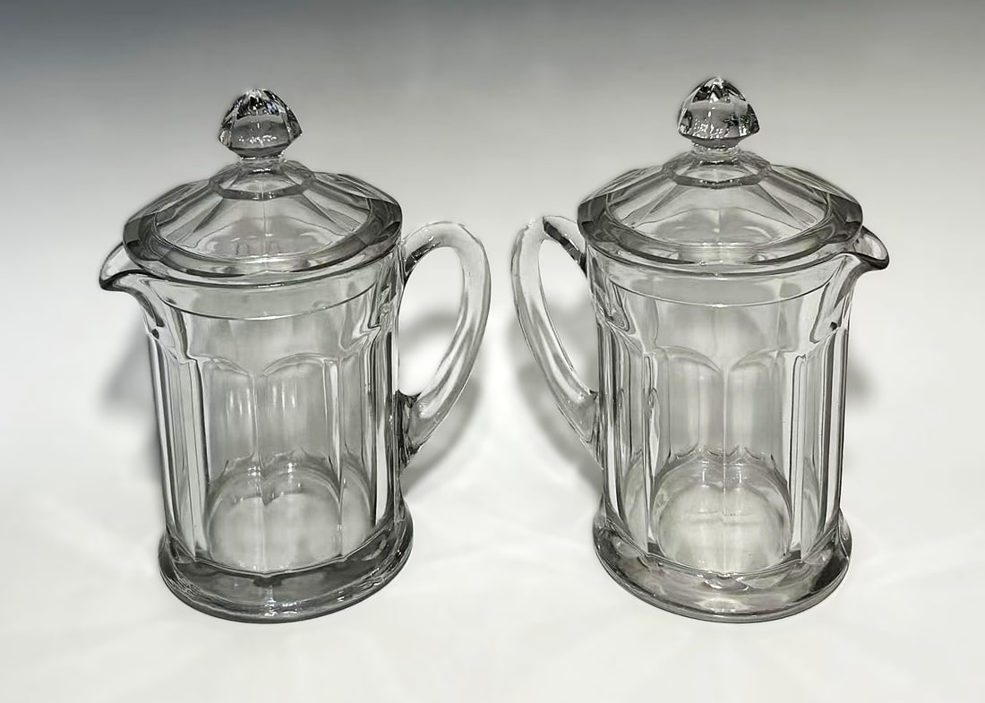 PAIR OF EARLY AMERICAN PATTERN GLASS ( EAPG) LIDDED JUGS: Circa 1910 to 1920s, each featuring a paneled body with applied handle, pouring spout, and fitted domed lid with faceted finial. The glass exhibits classic EAPG characteristics, including air bubble i