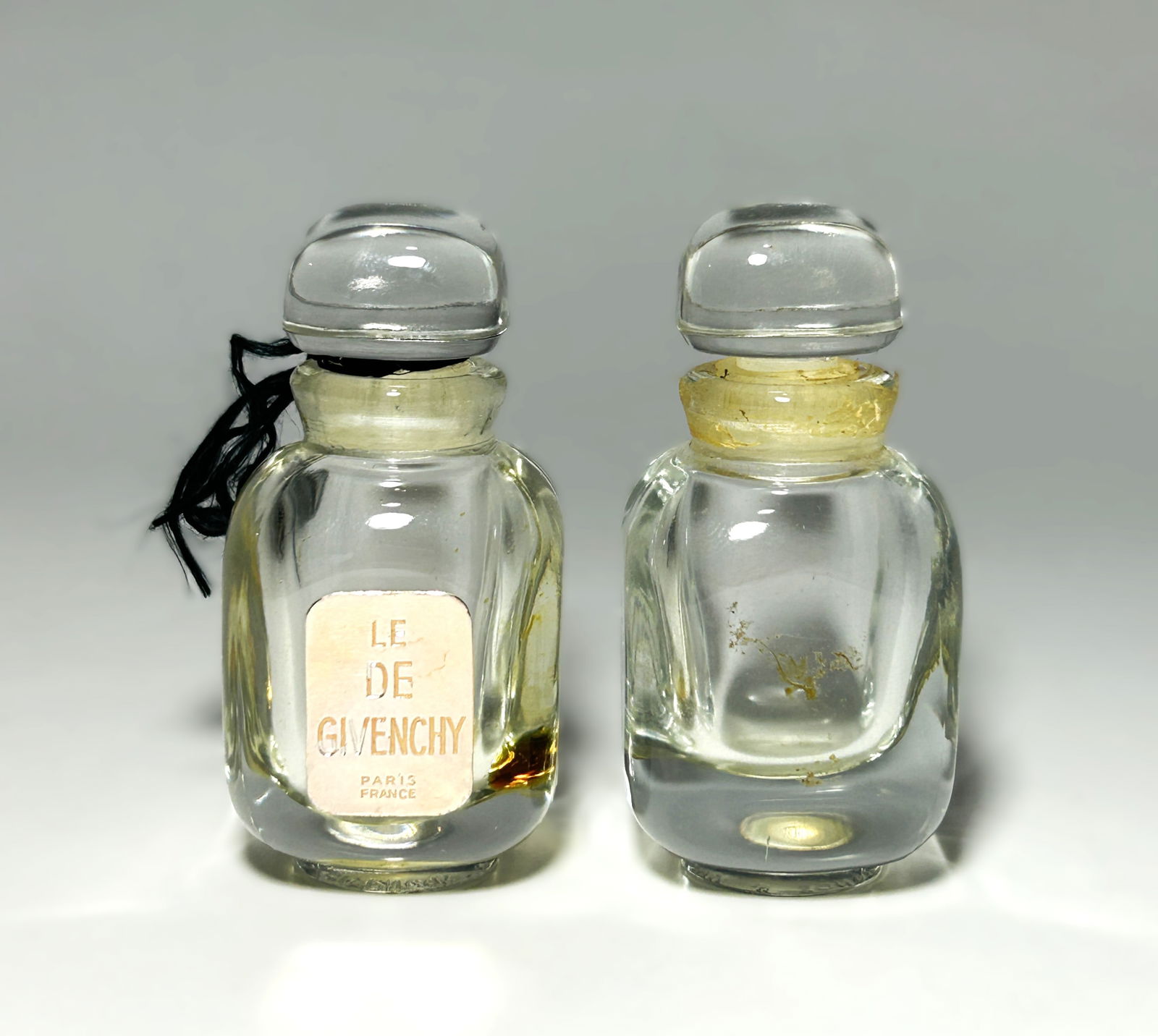TWO VINTAGE GIVENCHY “LE DE GIVENCHY?? PERFUME BOTTLES: Both of identical design, of clear molded glass with a rounded rectangular body and heavy base, fitted with matching clear glass stoppers of flattened domed form. One bottle retains its original front