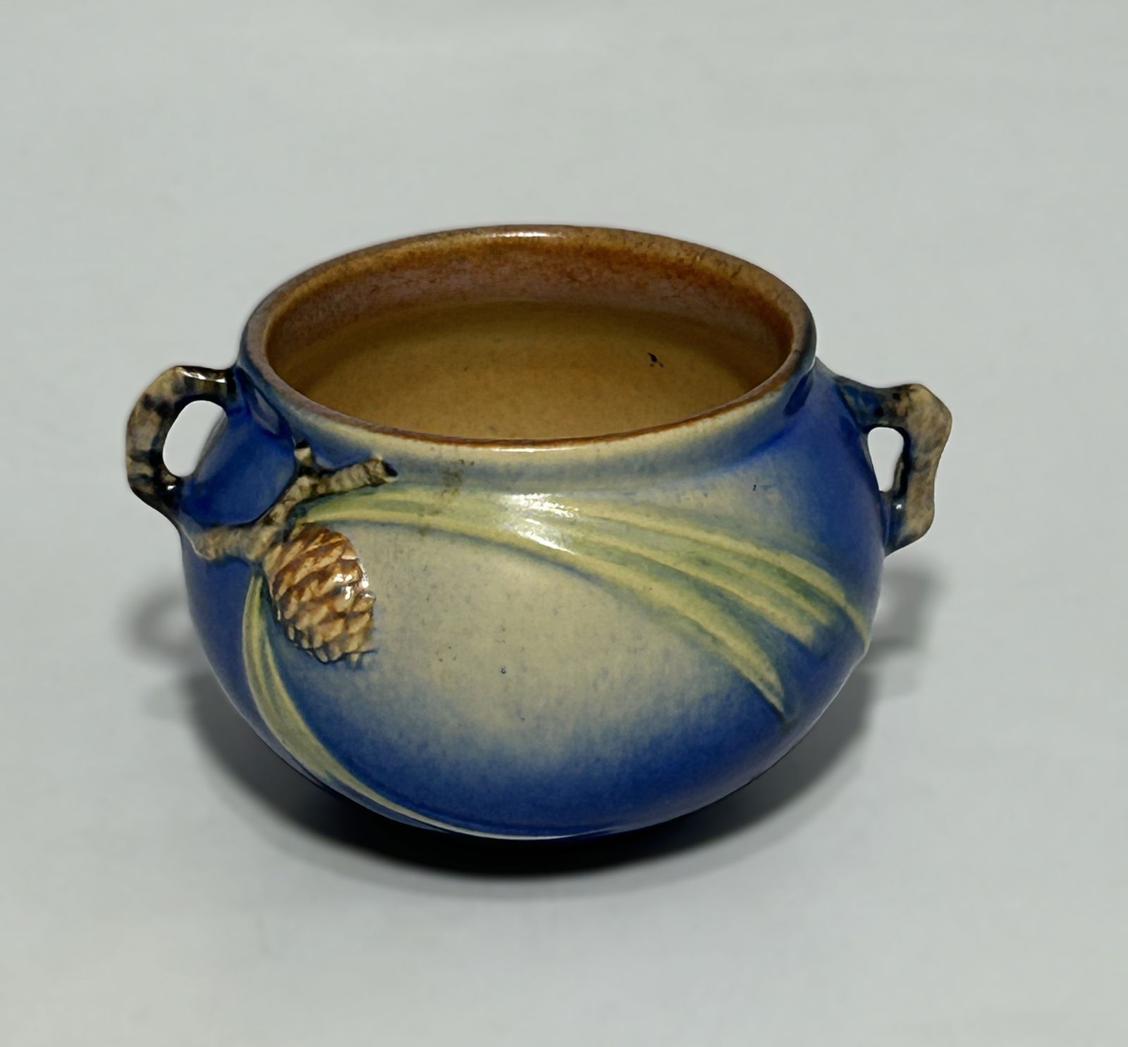 ROSEVILLE POTTERY "BLUE PINE CONE" JARDINIÈRE, CIRCA 1930S: The jardinière features a rounded, bulbous form with two opposing pierced lug handles. Finished in a rich matte blue glaze, the exterior is decorated with raised relief pine cones, needles, and sweep