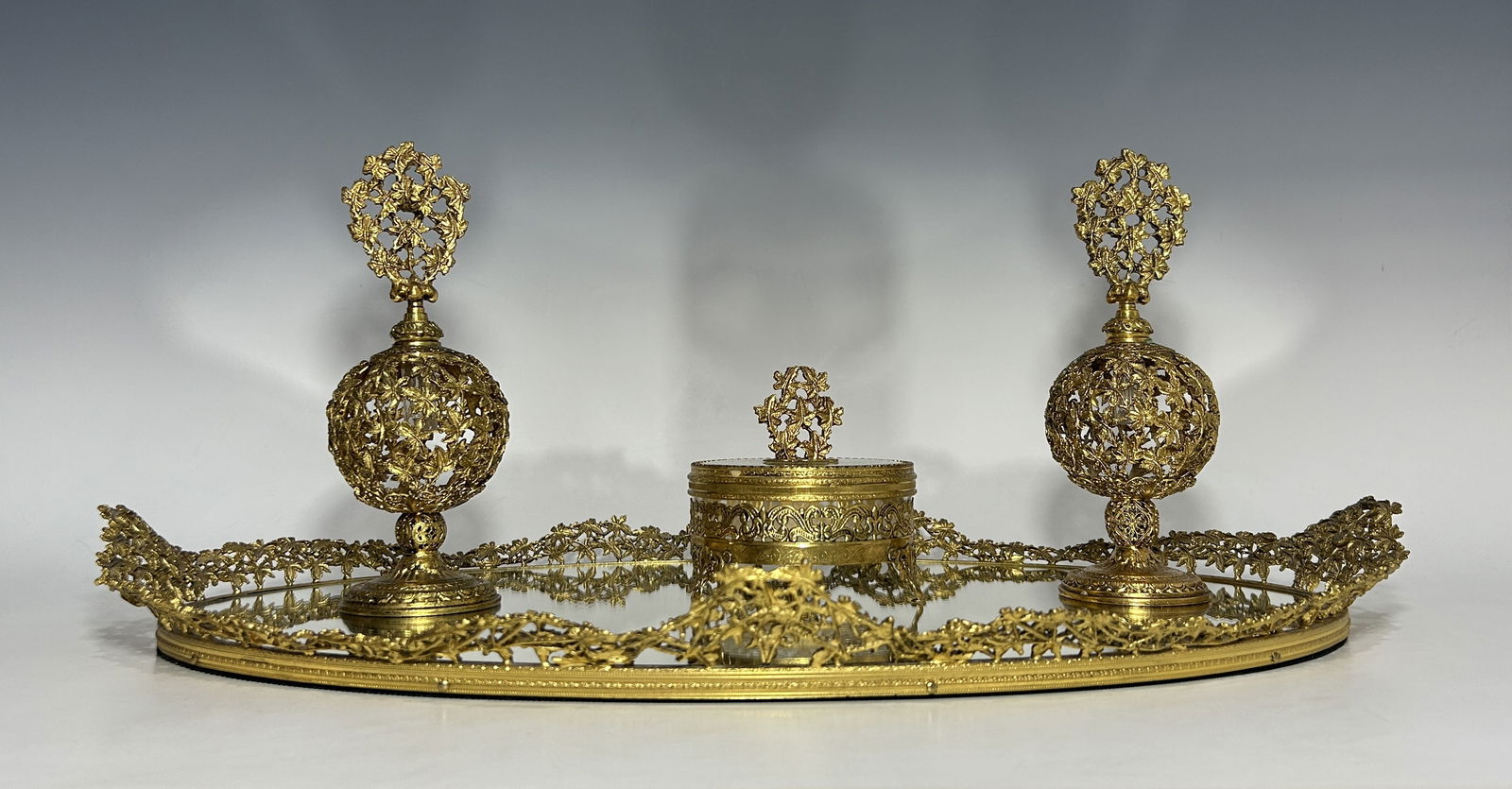 VINTAGE ORMOLU VANITY MIRROR TRAY WITH PERFUME BOTTLES AND TRINKET BOX: Decorative mid- century gilt-metal vanity set including an oval mirrored tray with openwork foliate gallery, a pair of matching ormolu perfume bottles with pierced spherical bodies and foliate stopper