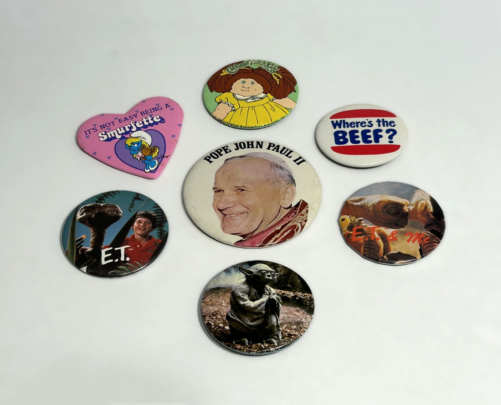 GROUP OF 7 VINTAGE POP CULTURE AND ADVERTISING PINBACK BUTTONS: including examples featuring Pope John Paul II, E.T., Yoda from Star Wars, Cabbage Patch Kids, Smurfette, and the advertising slogan “Where’s the Beef?”. Also includes an E.T. & Me button. M