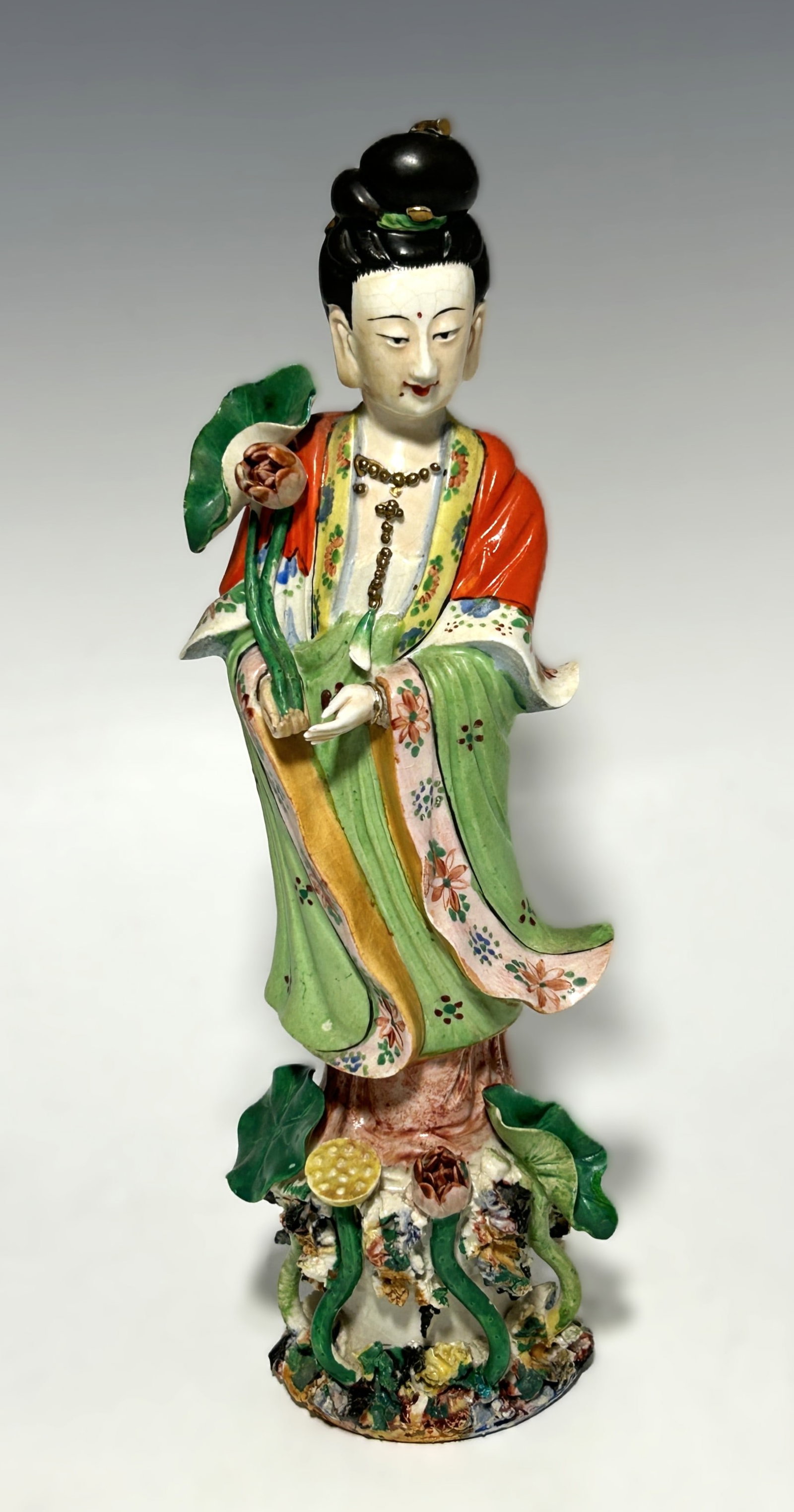 CHINESE FAMILLE ROSE PORCELAIN GUANYIN FIGURINE: Early 20th century, featuring the goddess with a serene expression and downcast eyes, holding a long-stemmed lotus flower in her right hand. She is adorned in traditional flowing robes of green, yello