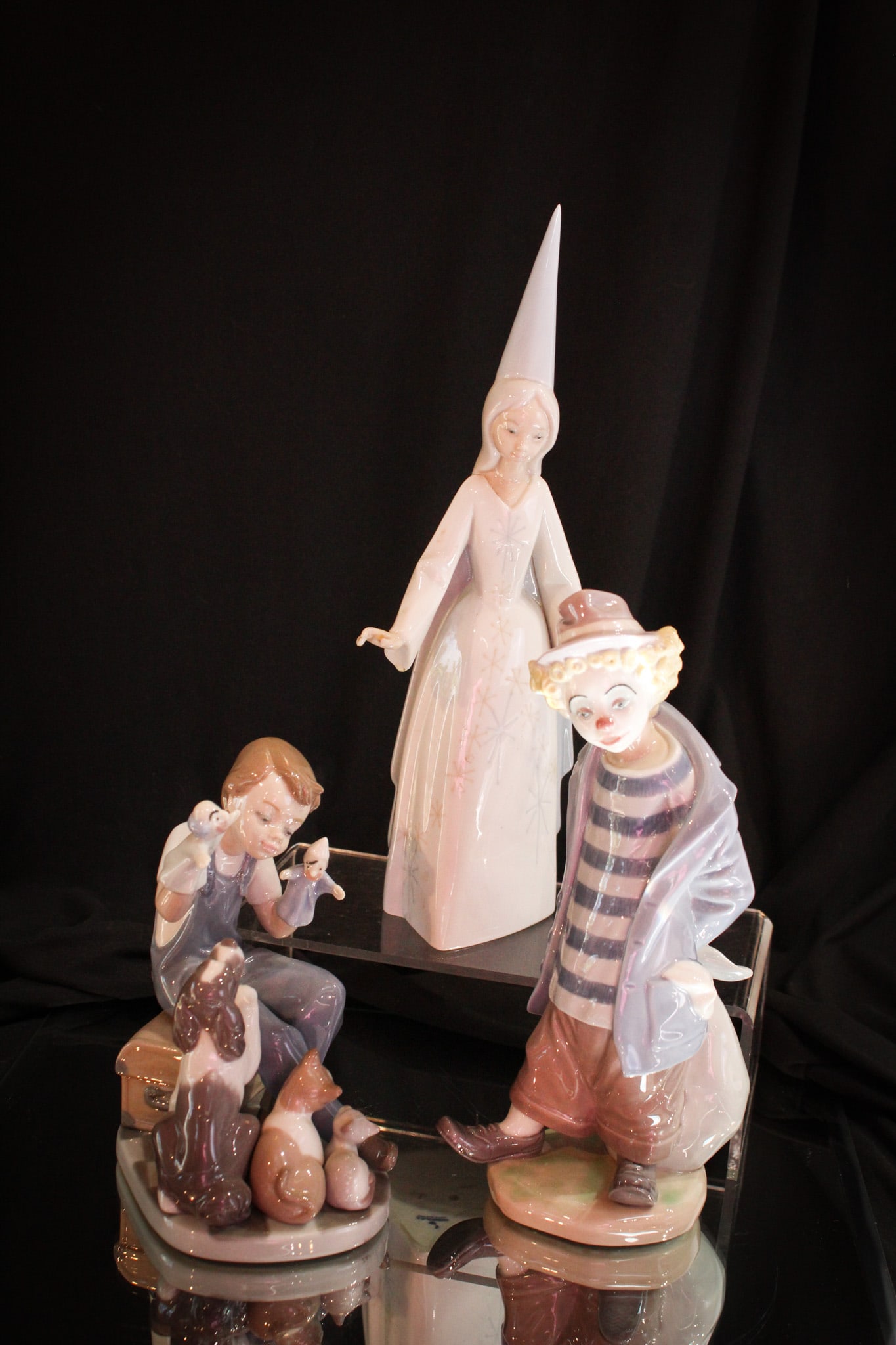 3 pc Lot Lladro Porcelain Figurines: Nice array of figures in this bountiful lot of signed Lladro porcelain figurines. Lot is comprised of a child clown, a boy playing with puppets, and a girl wizard or godmother. Tallest measures 11".