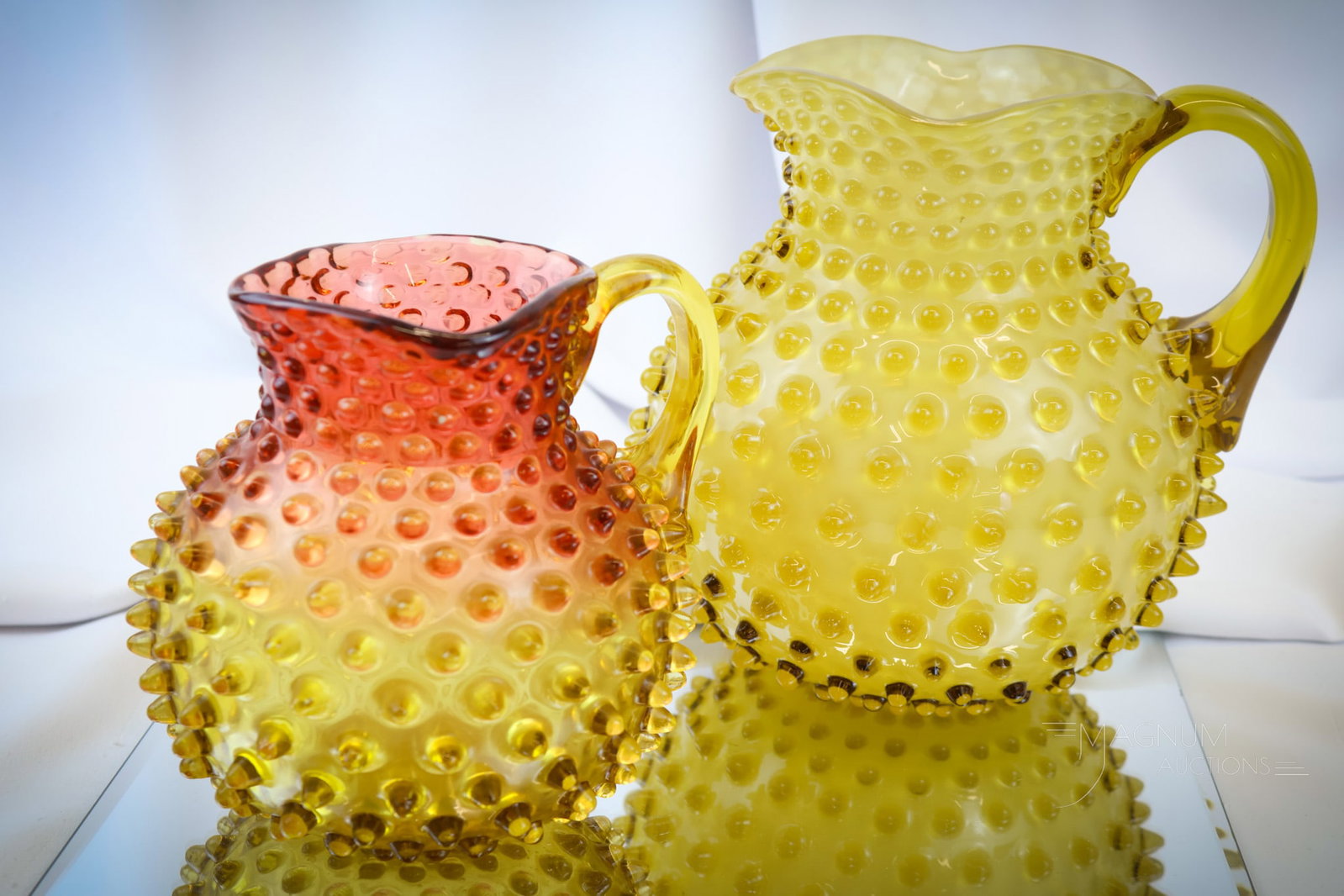 2 pc Lot Hobbs Hobnail Amberina & Cased Victorian Glass Pitchers: Two delightful and hard-to-find pitchers made by Hobbs. The first is an amberina hobnail milk-size pitcher. The second is a cased amber glass hobnail water pitcher. The tallest measures 7 1/2"". The a