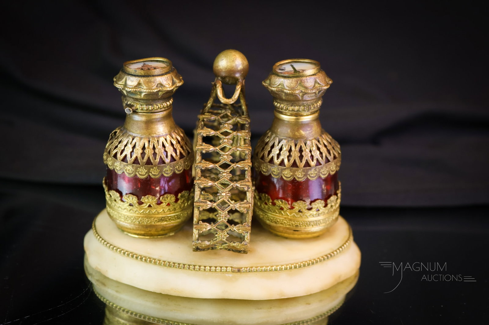 Parisian Grand Tour Double Cranberry Perfume Set: A select find for your collection. This is a Grand Tour double perfume set. It features two cranberry bottles with Parisian scenes in the tops. The brass frame resides on a marble base. The set measur
